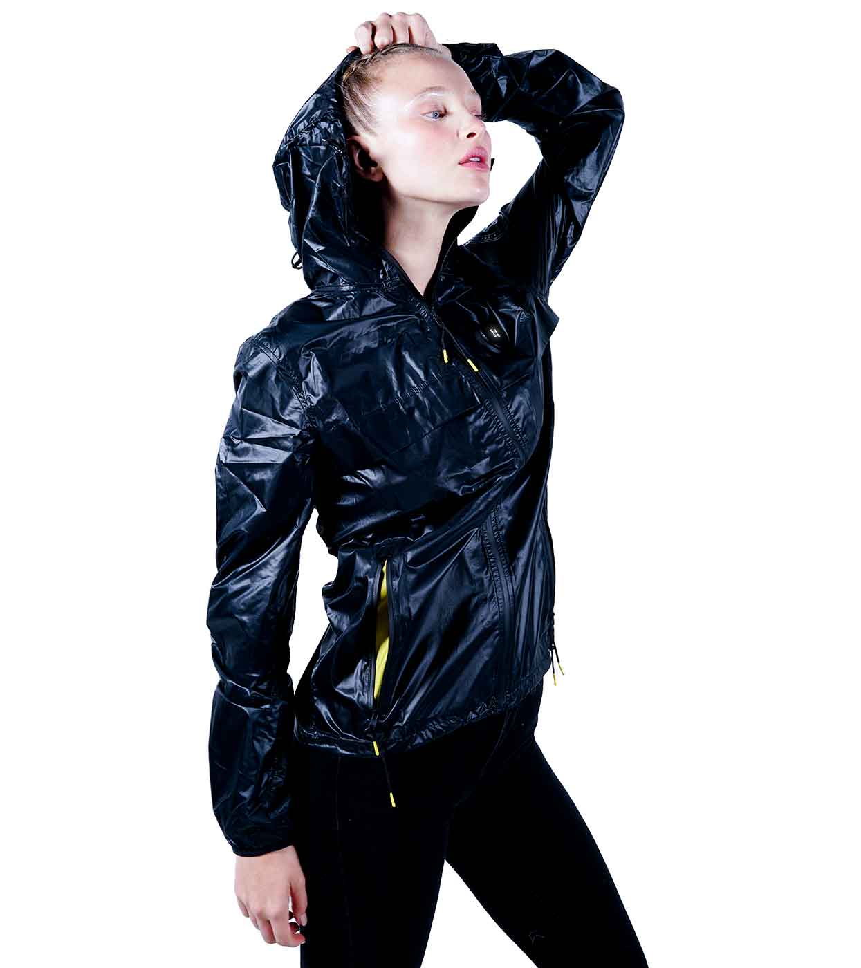 A person is wearing the BLACK WINDBREAKER by Machina - Wearable Technology, featuring a shiny black finish with striking yellow zipper accents. They are posing stylishly with one hand on their head, making this ultralight nylon jacket stand out against the white background.