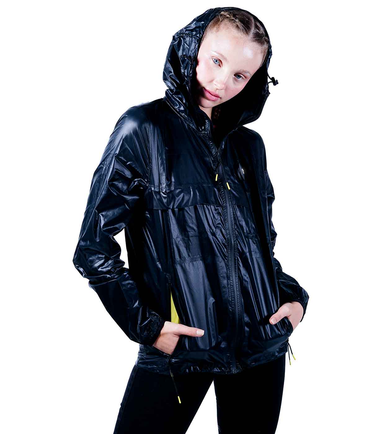 A person wearing the BLACK WINDBREAKER by Machina - Wearable Technology stands against a plain white background. This shiny black hooded jacket, featuring sleek smart windbreaker technology, includes a zipper and striking yellow accents. Styled with black pants and light-colored braided hair, it epitomizes seamless fashion innovation in design.