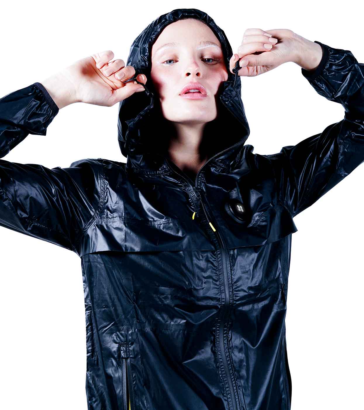 A person models the BLACK WINDBREAKER by Machina - Wearable Technology, crafted from ultralight nylon with a glossy finish. The shiny black jacket, complete with a zipper, is set against a white background as the individual strikes a pose with hands near the head. They gaze upward slightly, their hair neatly tucked into the hood of this sleek windbreaker.