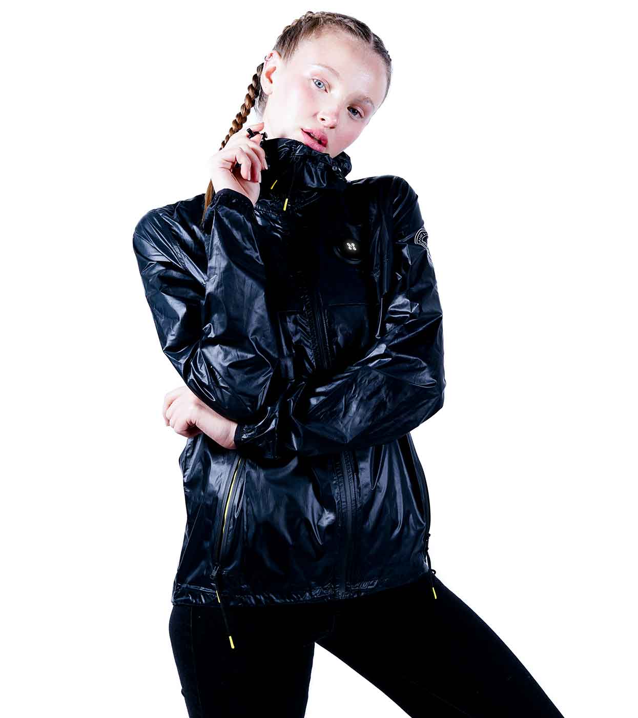 Against a white background, an individual with braided hair stands wearing the BLACK WINDBREAKER by Machina - Wearable Technology. Crafted from ultralight nylon, this shiny black jacket features a hood and proudly displays the logo on the chest. Holding the collar close to their mouth, they maintain a calm expression.