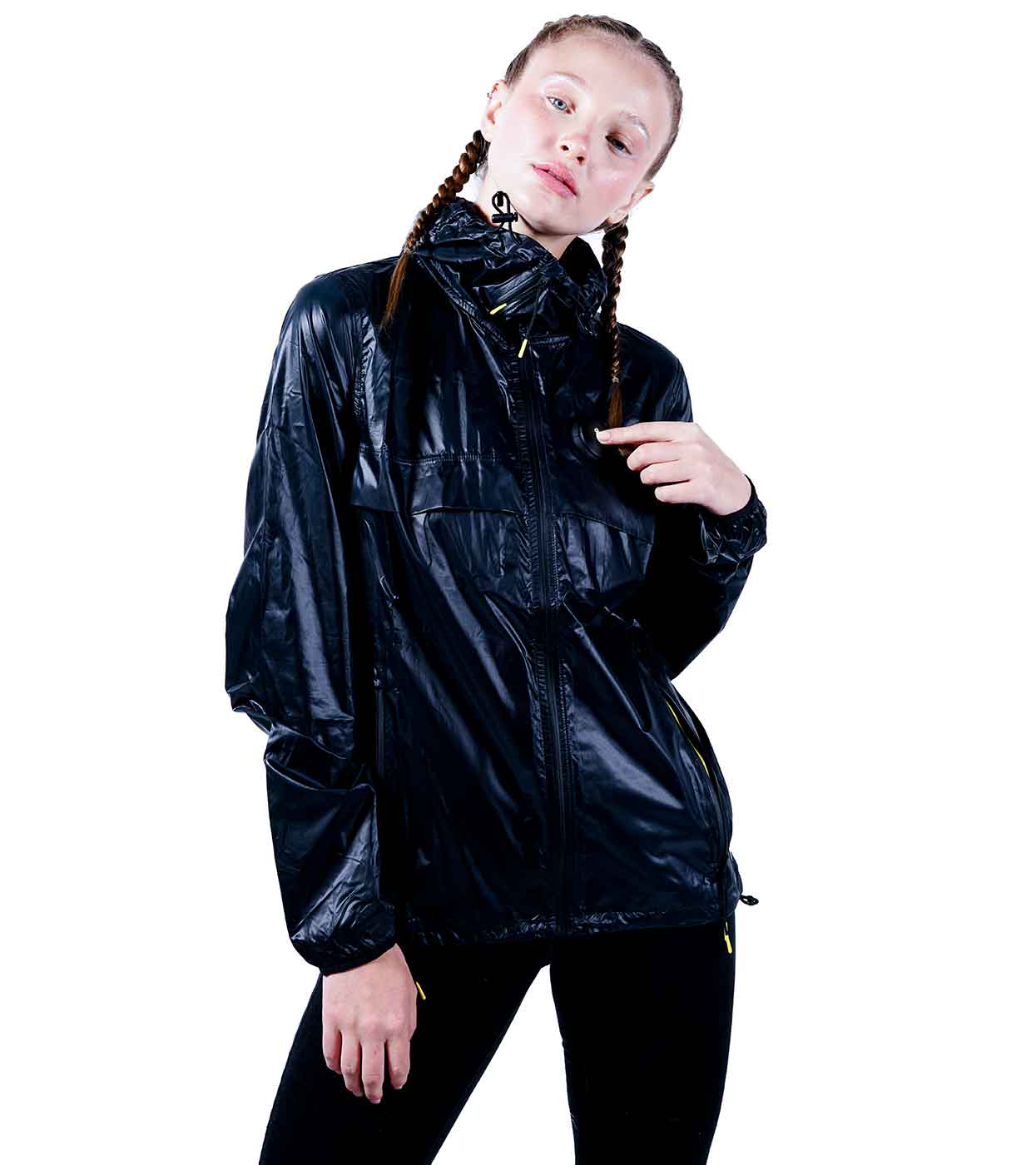 A person wearing the BLACK WINDBREAKER from Machina - Wearable Technology pairs it with black pants as they stand against a white background. Their hair is styled in two braids, adding flair to the ultralight nylon ensemble. They pose confidently, one hand on their hip and the other slightly raised at their side.