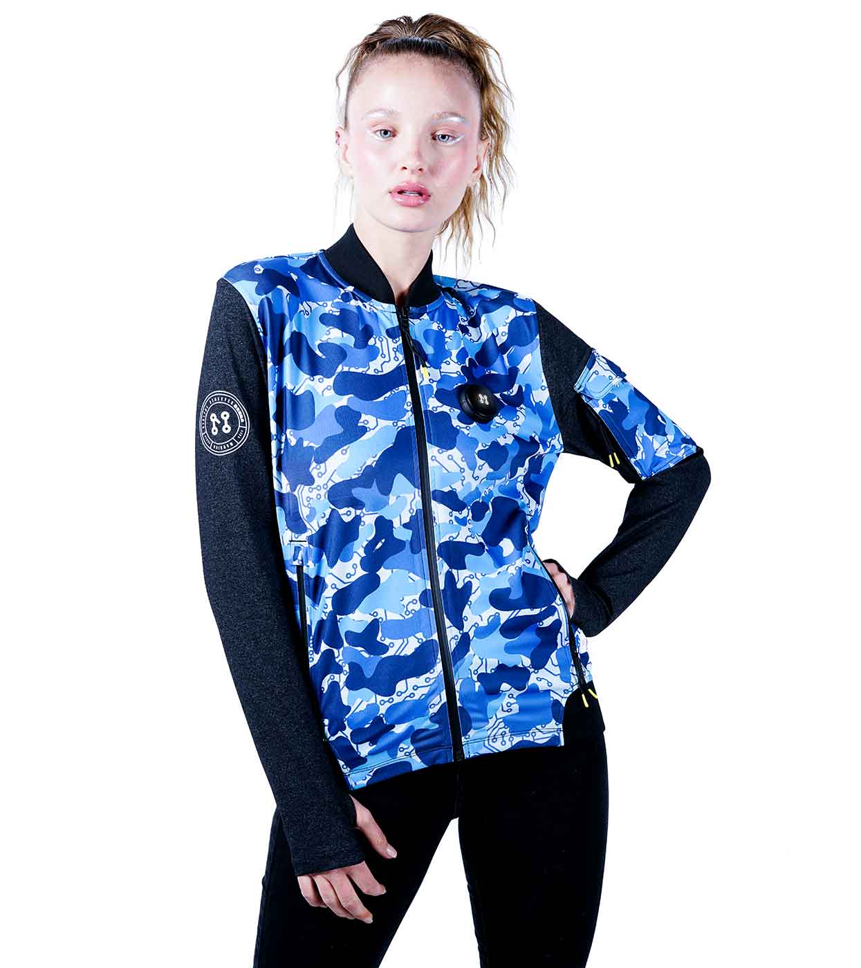 In front of a white backdrop, an individual confidently stands with one hand on their hip, showcasing the BLUE-CAMO BOMBER JACKET by Machina - Wearable Technology. The blue and white bomber jacket, featuring black sleeves and made from sustainable fabrics, complements their black pants perfectly.
