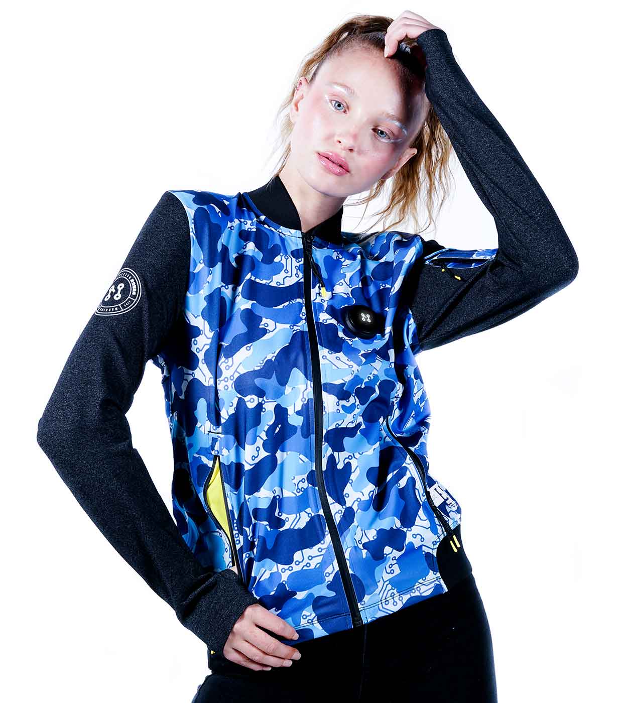 A person with long blonde hair is wearing the Machina - Wearable Technology BLUE-CAMO BOMBER JACKET, featuring a blue and white camouflage pattern with black sleeves. They are posing with one hand on their head against a white background.