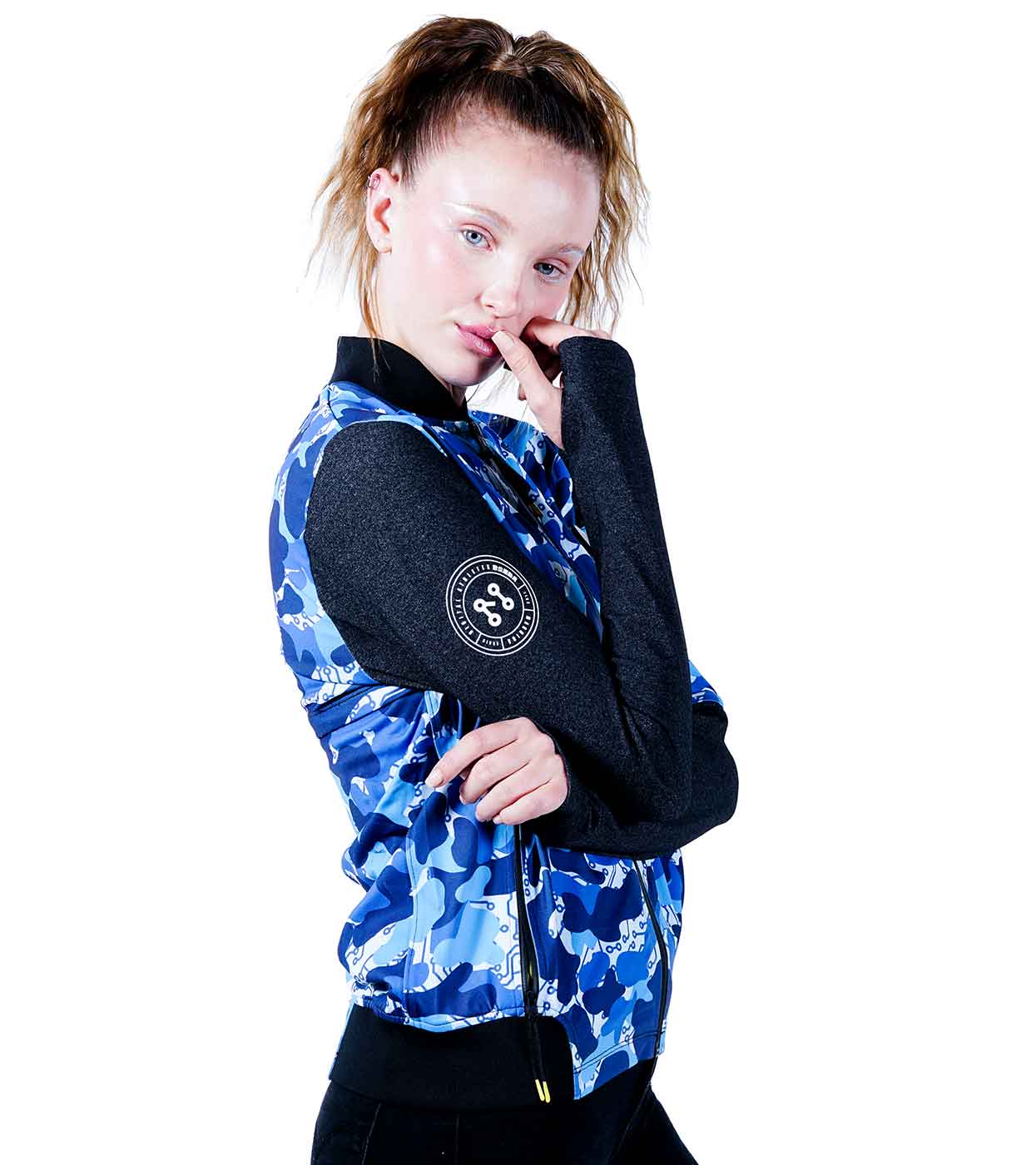 A woman poses confidently, one hand on her chin, wearing a stylish BLUE-CAMO BOMBER JACKET by Machina - Wearable Technology. Featuring a blue camouflage design with a black collar and sleeves, along with a distinctive circular logo, the outfit highlights her fashion-forward flair. Her hair is styled in a ponytail against a white background, embodying the spirit of fashion tech.