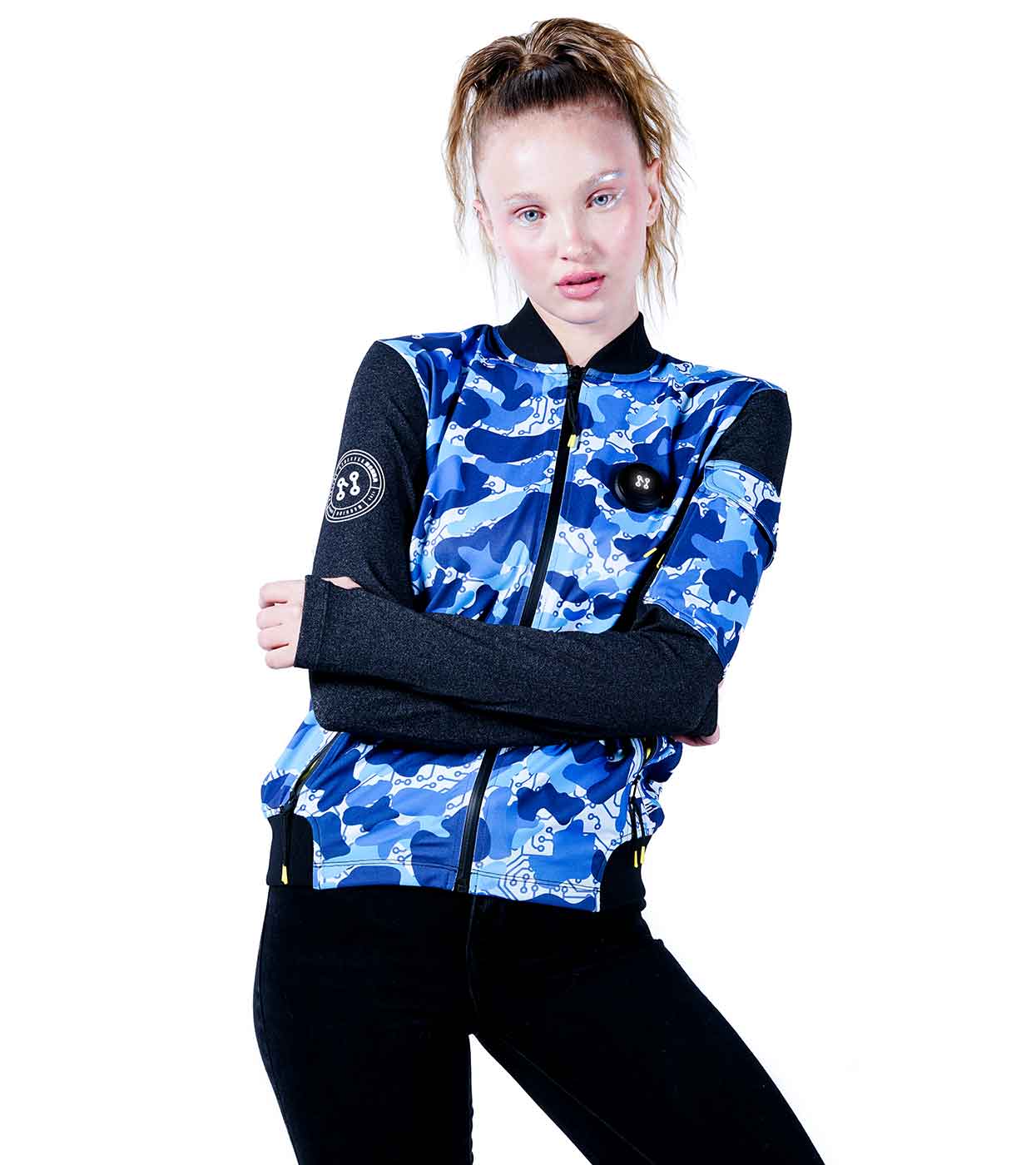 A person stands confidently with arms crossed, wearing the BLUE-CAMO BOMBER JACKET by Machina - Wearable Technology. They pair it with black sleeves and black pants, their light hair pulled back in a high ponytail against a plain white background.