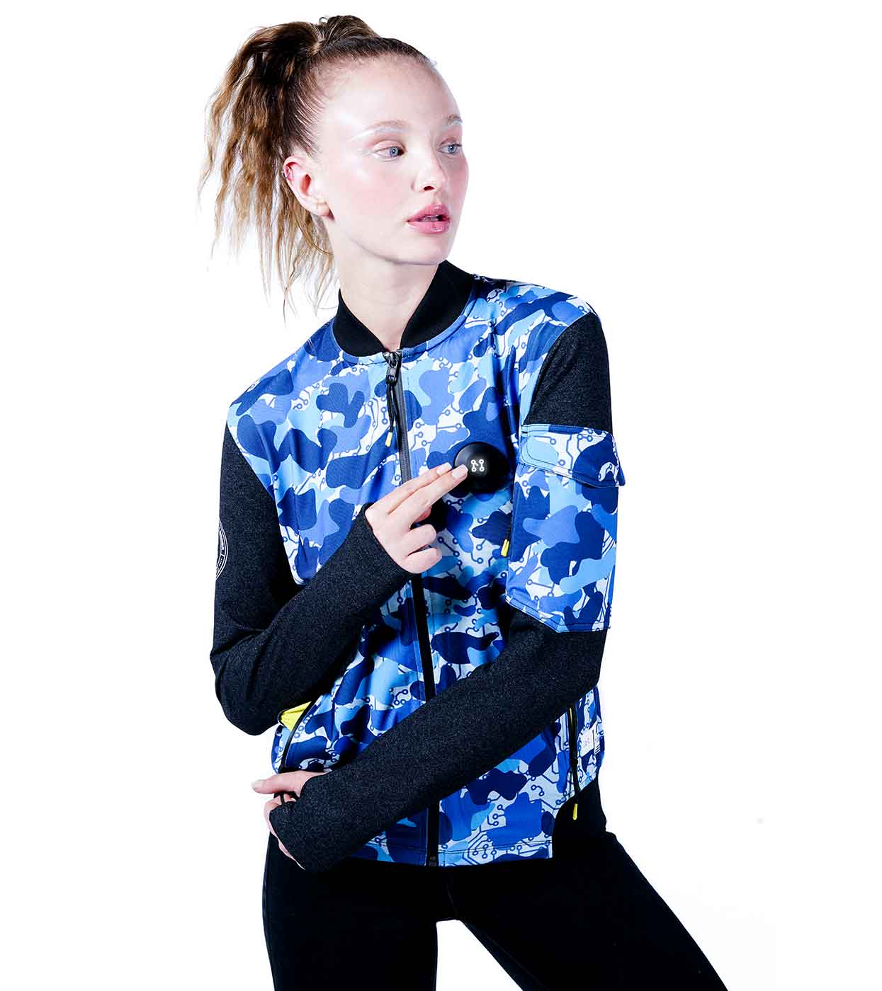 A person dressed in the BLUE-CAMO BOMBER JACKET by Machina - Wearable Technology, featuring a striking blue camo design and black sleeves, points at a badge on the jacket. The individual, highlighting innovative fashion tech, has long hair tied up and gazes to the side against a plain white background.