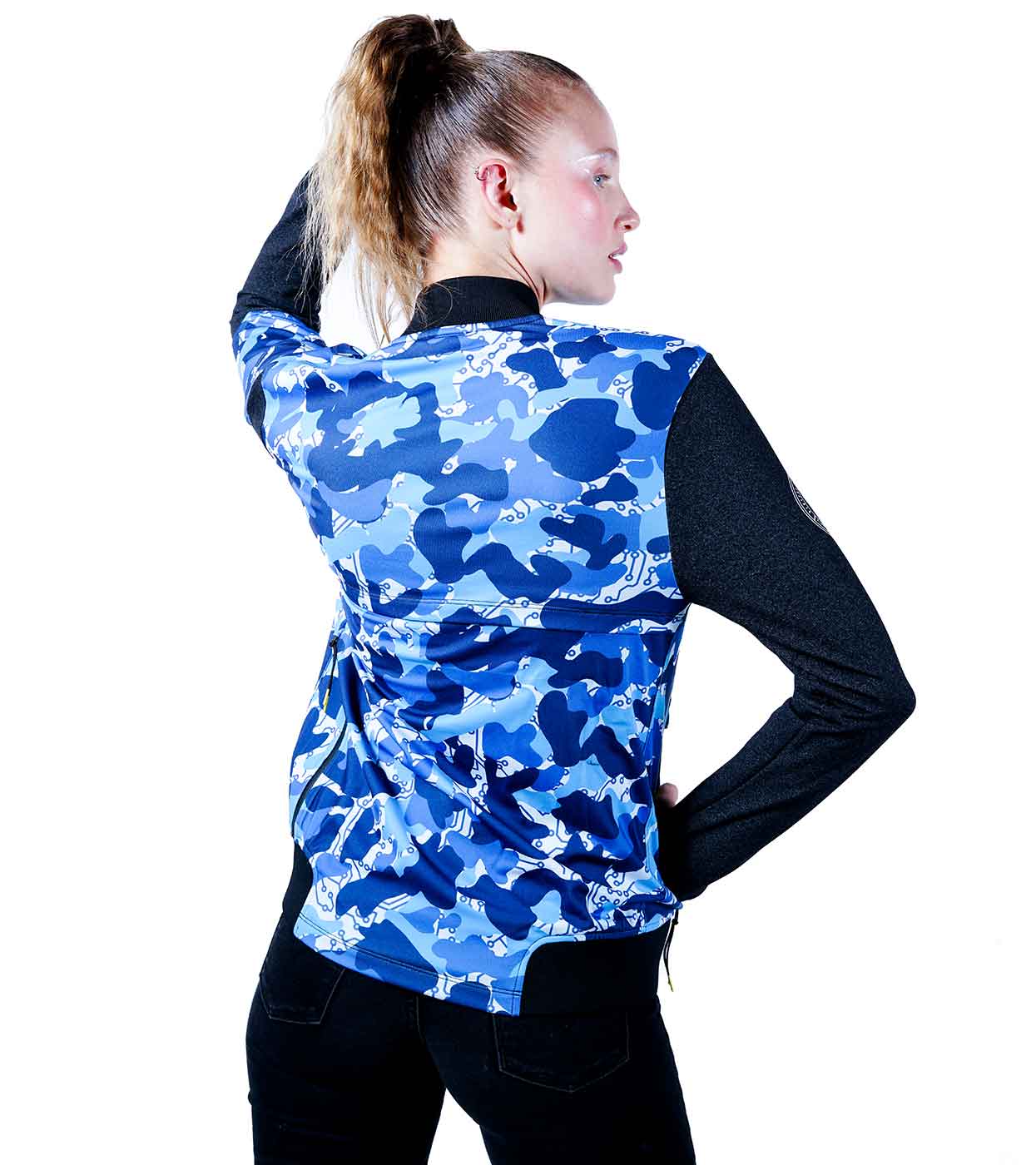 A person with a ponytail, facing away from the camera, is stylishly dressed in Machina's BLUE-CAMO BOMBER JACKET, featuring an innovative blend of fashion technology and sleek design with its blue and white body and black sleeves. They confidently pose with one hand on their hip and the other behind their head.