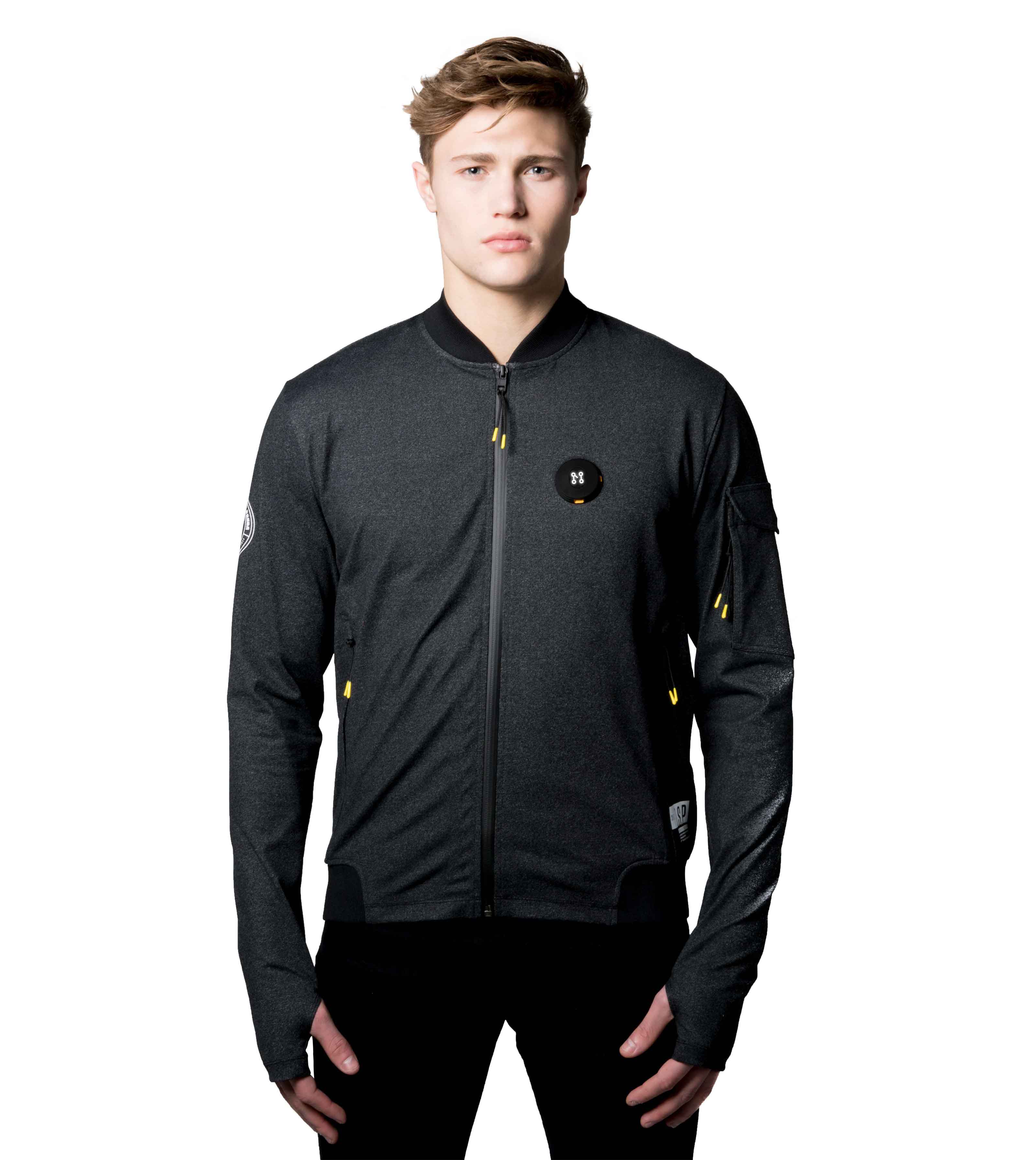 An individual is dressed in a BLACK BOMBER JACKET by Machina - Wearable Technology, characterized by a small logo on the chest and a patch on the sleeve. This jacket, crafted from sustainable fabrics, includes thumb holes in the sleeves and is displayed against a plain white background.