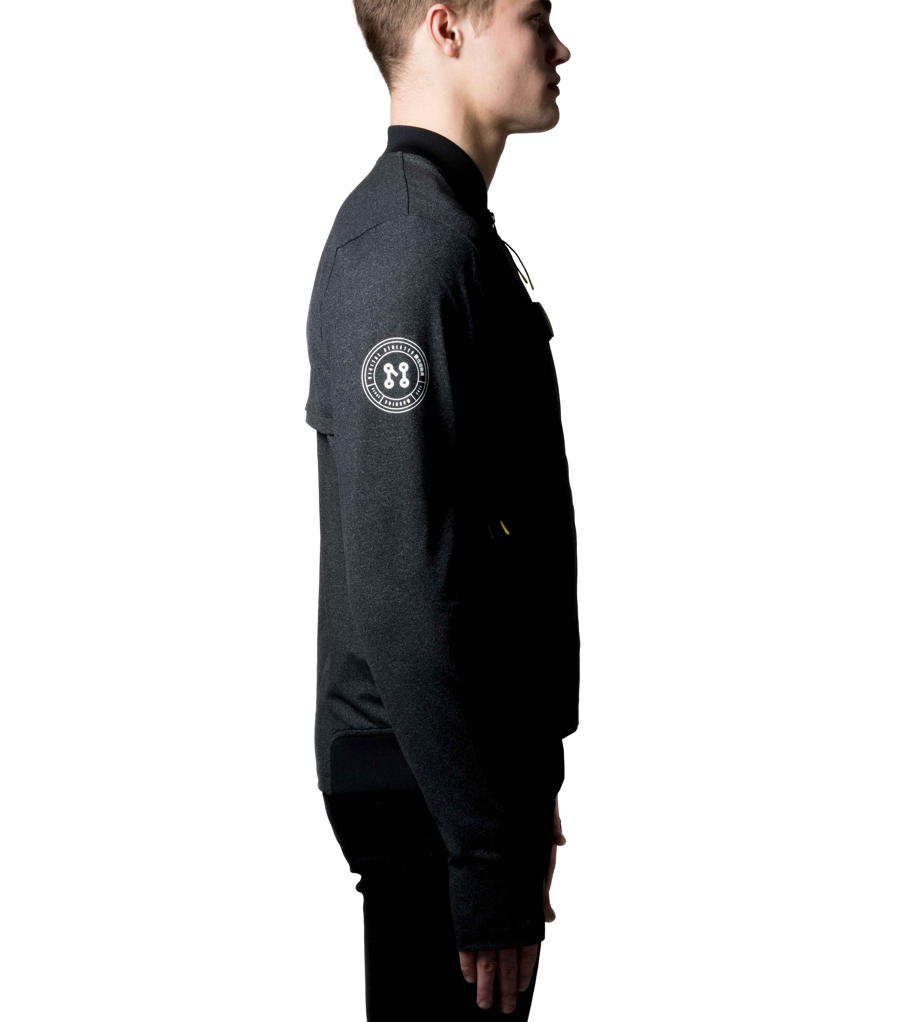 A person is posed in profile against a white background, wearing the BLACK BOMBER JACKET by Machina - Wearable Technology. This smart, minimalist, and modern jacket is crafted from sustainable fabrics and features a distinctive circular patch with a symmetrical design on the sleeve.