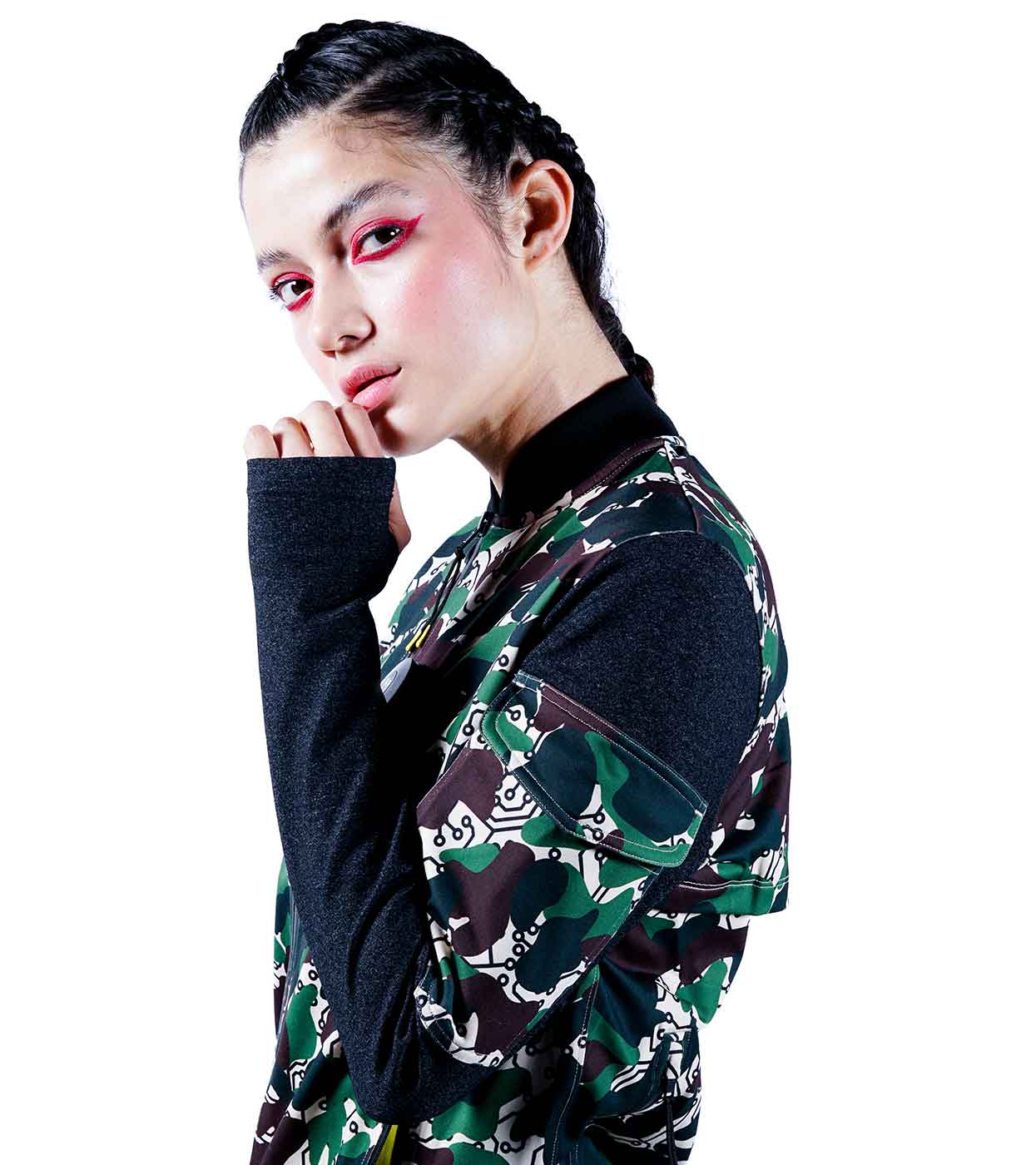 A person with braided hair, wearing the DESIERTO CAMO BOMBER JACKET by Machina - Wearable Technology, poses with a contemplative expression. Bold pink eye makeup accentuates their look against a plain white background.