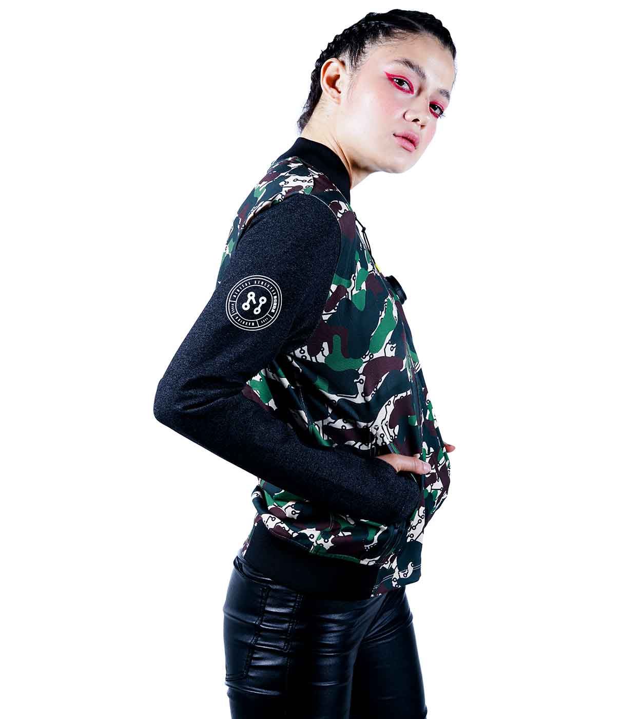 A person with braided hair and bold makeup poses confidently in the Machina - Wearable Technology DESIERTO CAMO BOMBER JACKET, which features black sleeves and a circular patch on the arm. Paired with black leather pants, they exude confidence as they glance to the side. The plain white background accentuates their edgy style.