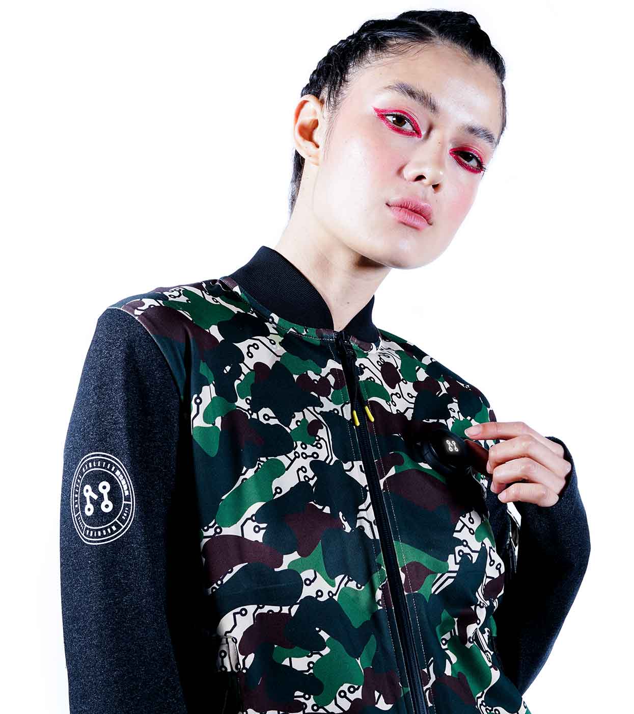A person with braided hair wears the DESIERTO CAMO BOMBER JACKET from Machina - Wearable Technology, which features a circular patch on the sleeve. With bold red eye makeup and standing against a plain white background, their look merges classic style with the innovation of fashion tech.