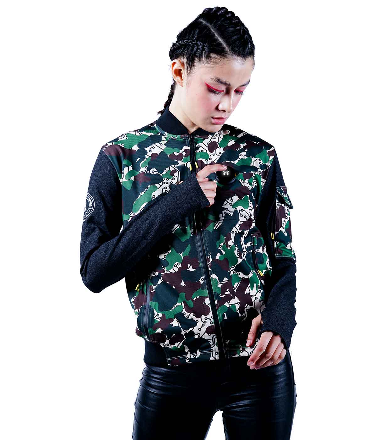 A person with braided hair stands against a white background, wearing the DESIERTO CAMO BOMBER JACKET by Machina - Wearable Technology, crafted from sustainable fabrics. The black sleeves complement their black pants as they hold and examine a small object in their hand.