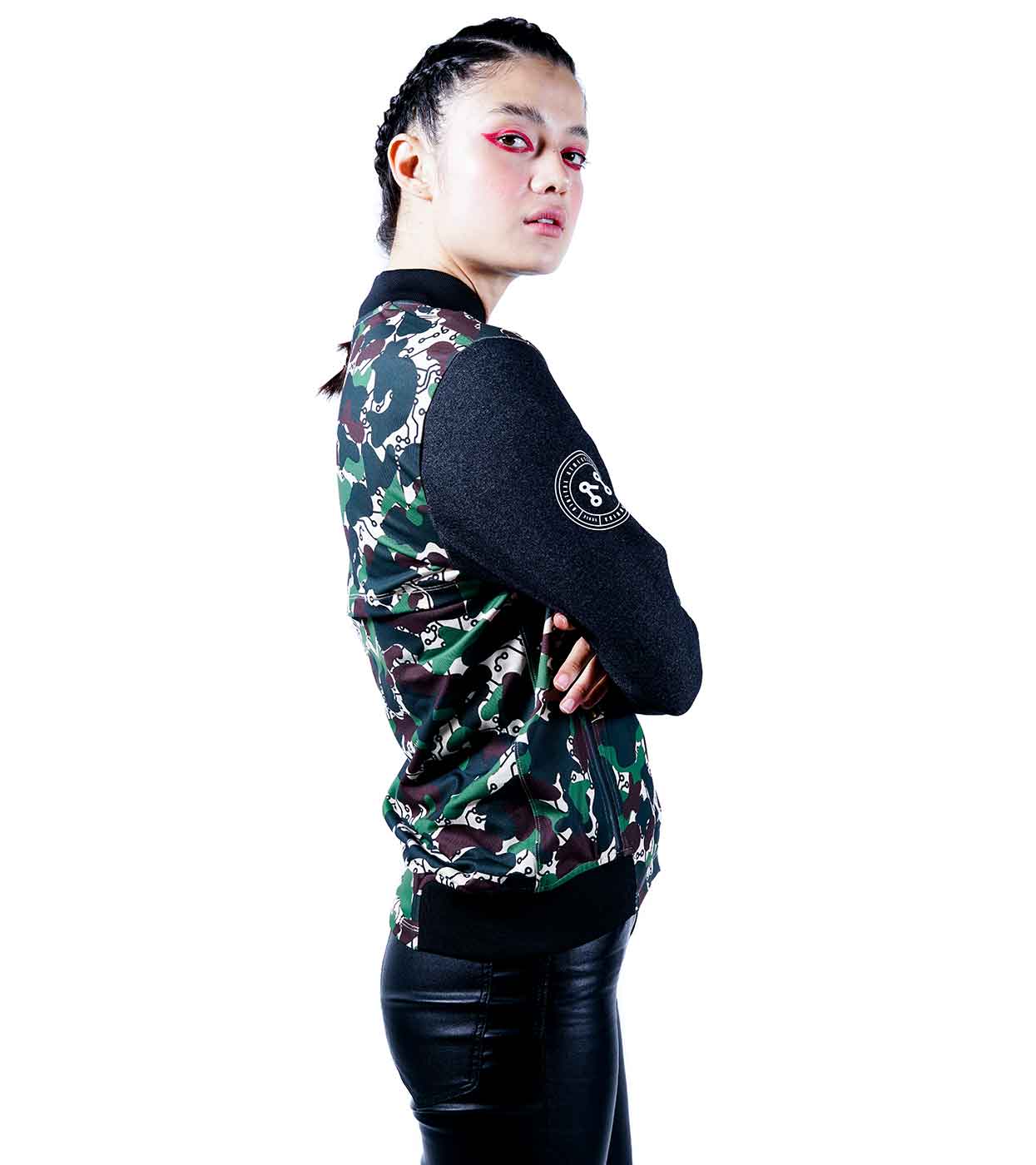 With braided hair, red eye makeup, and crossed arms, the individual exudes confidence as they glance over their shoulder. They are wearing the stylish DESIERTO CAMO BOMBER JACKET by Machina - Wearable Technology, paired with black-sleeved leather pants. The white background enhances their ability to seamlessly blend style with cutting-edge fashion tech.