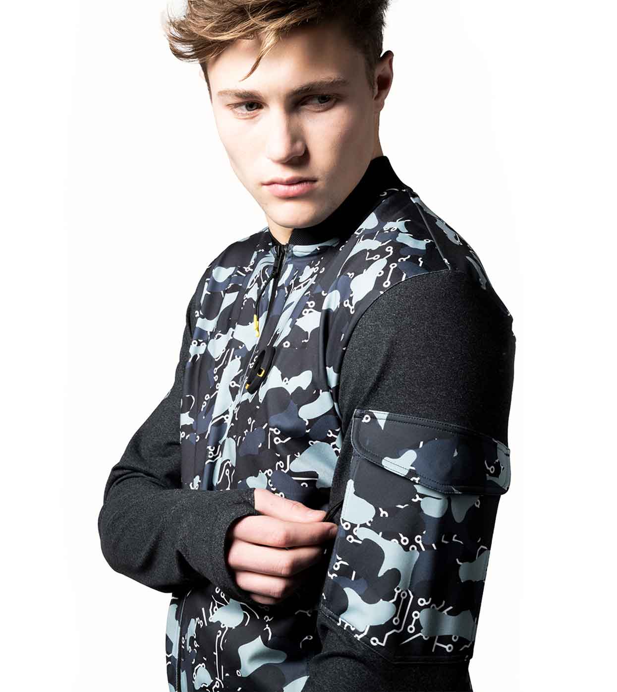 A person wearing the BLACK-CAMO BOMBER JACKET by Machina - Wearable Technology stands against a white background. The jacket showcases an abstract pattern in shades of gray and blue, complemented by its dark sleeves. The person gazes slightly downward, with one hand resting on the opposite sleeve.
