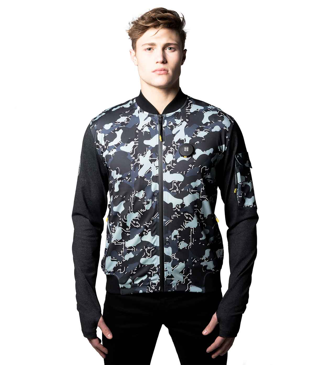 A person wearing the BLACK-CAMO BOMBER JACKET by Machina - Wearable Technology stands against a white background. They have short hair and are looking straight ahead with their hands in their pockets.