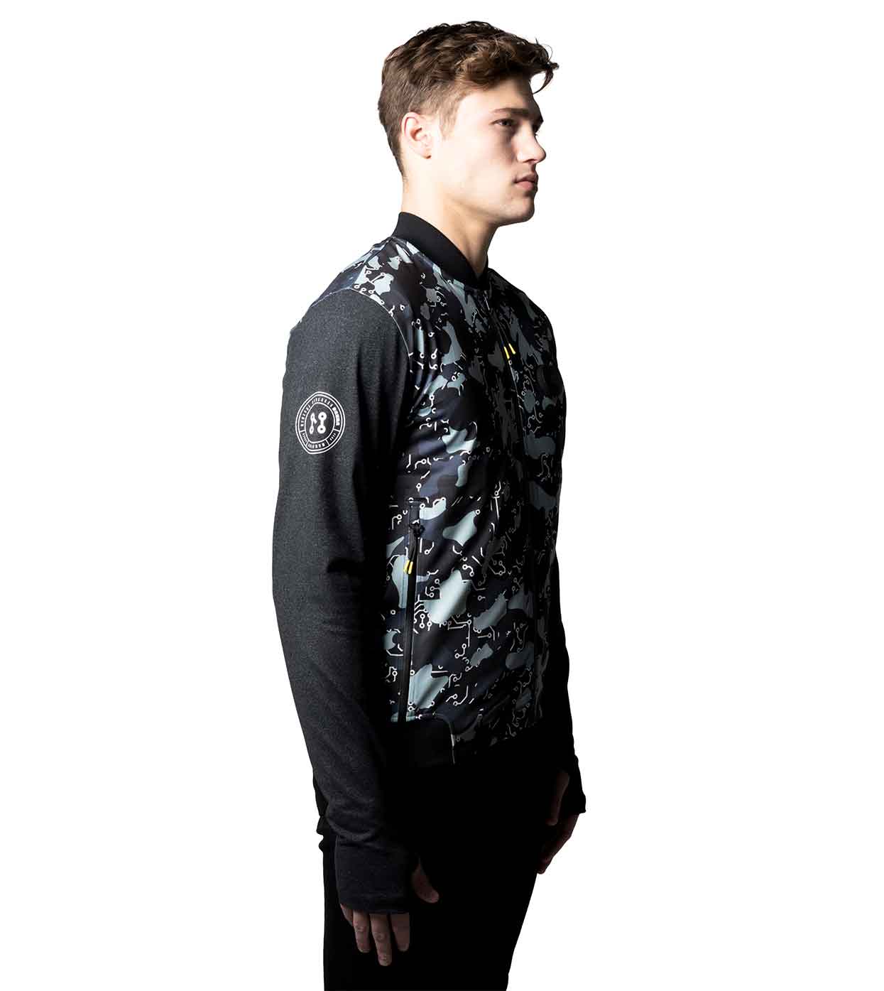 A man stands in profile against a white background, wearing a BLACK-CAMO BOMBER JACKET by Machina - Wearable Technology, featuring dark sleeves and a logo on the left sleeve. His hair is styled short, and he is looking straight ahead.