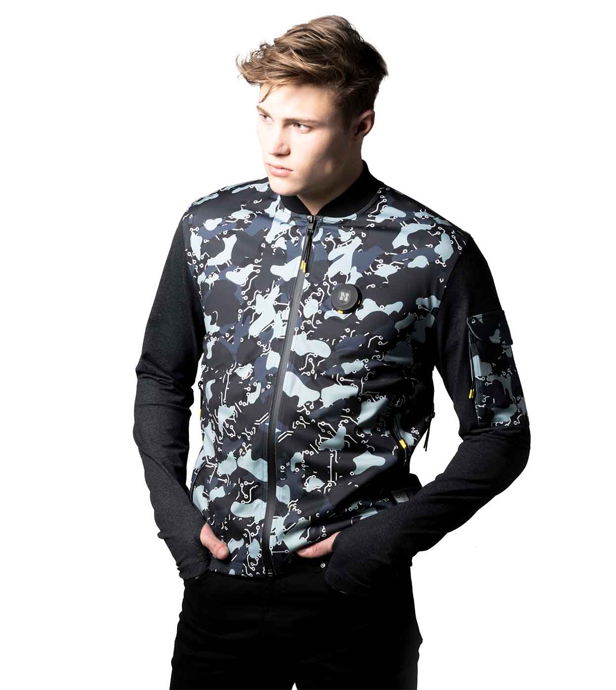 A young man wearing the BLACK-CAMO BOMBER JACKET from Machina - Wearable Technology, featuring a camo design and black sleeves, stands with his hands in his pockets against a white background.