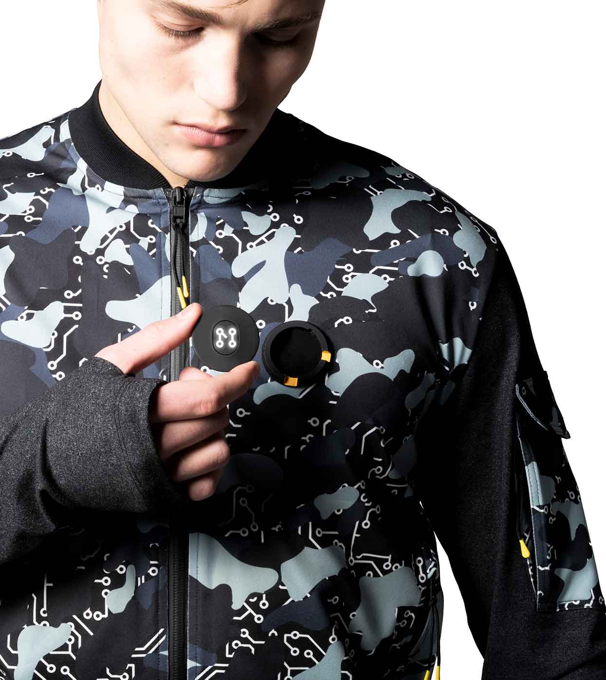 A person clad in the BLACK-CAMO BOMBER JACKET by Machina - Wearable Technology, featuring a high-tech design, looks down while holding a small round device. Set against a white background, their expression appears focused.