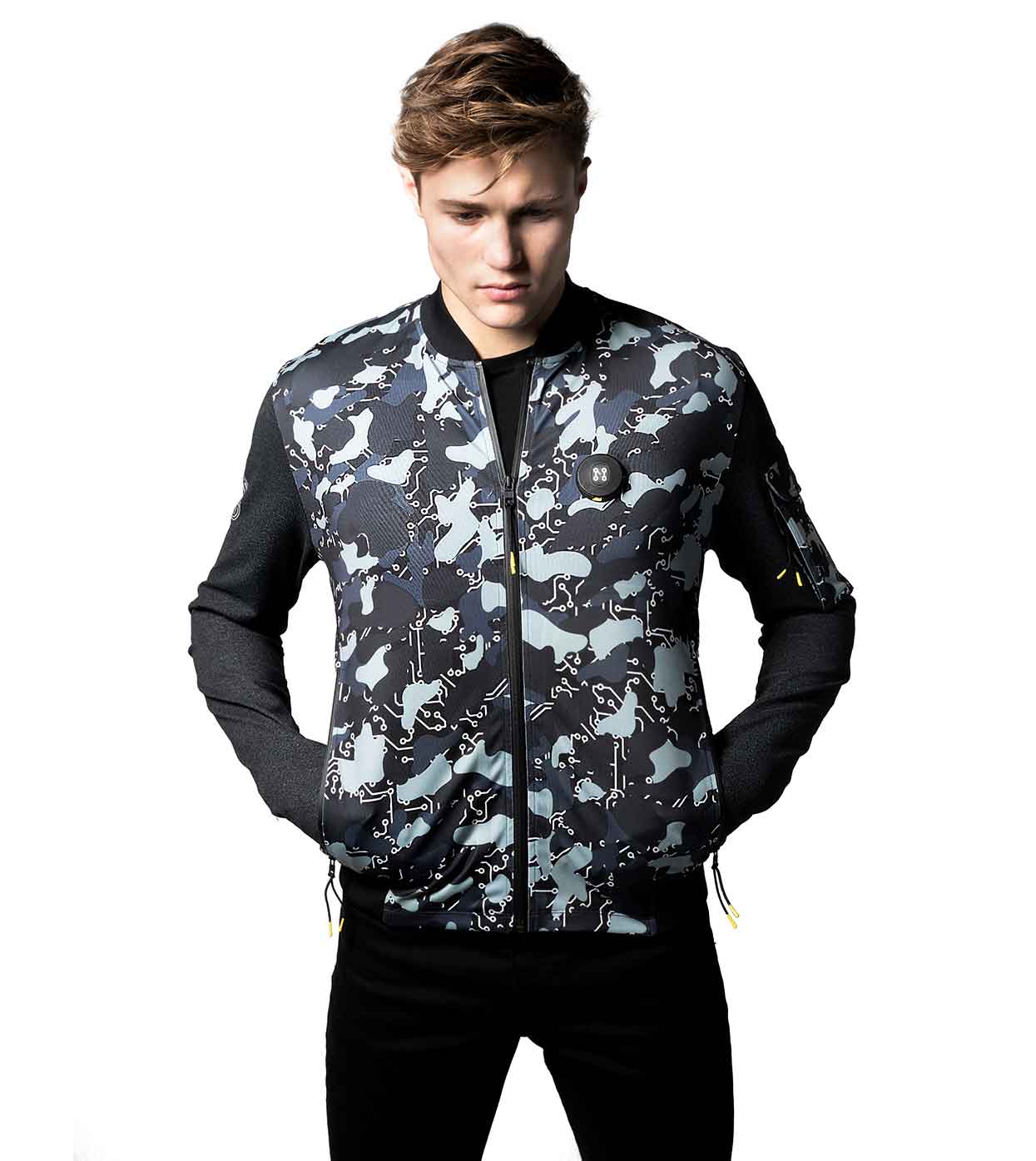 A person wearing the BLACK-CAMO BOMBER JACKET by Machina - Wearable Technology, featuring a camo pattern with dark sleeves, stands against a white background, looking down with hands in pockets.