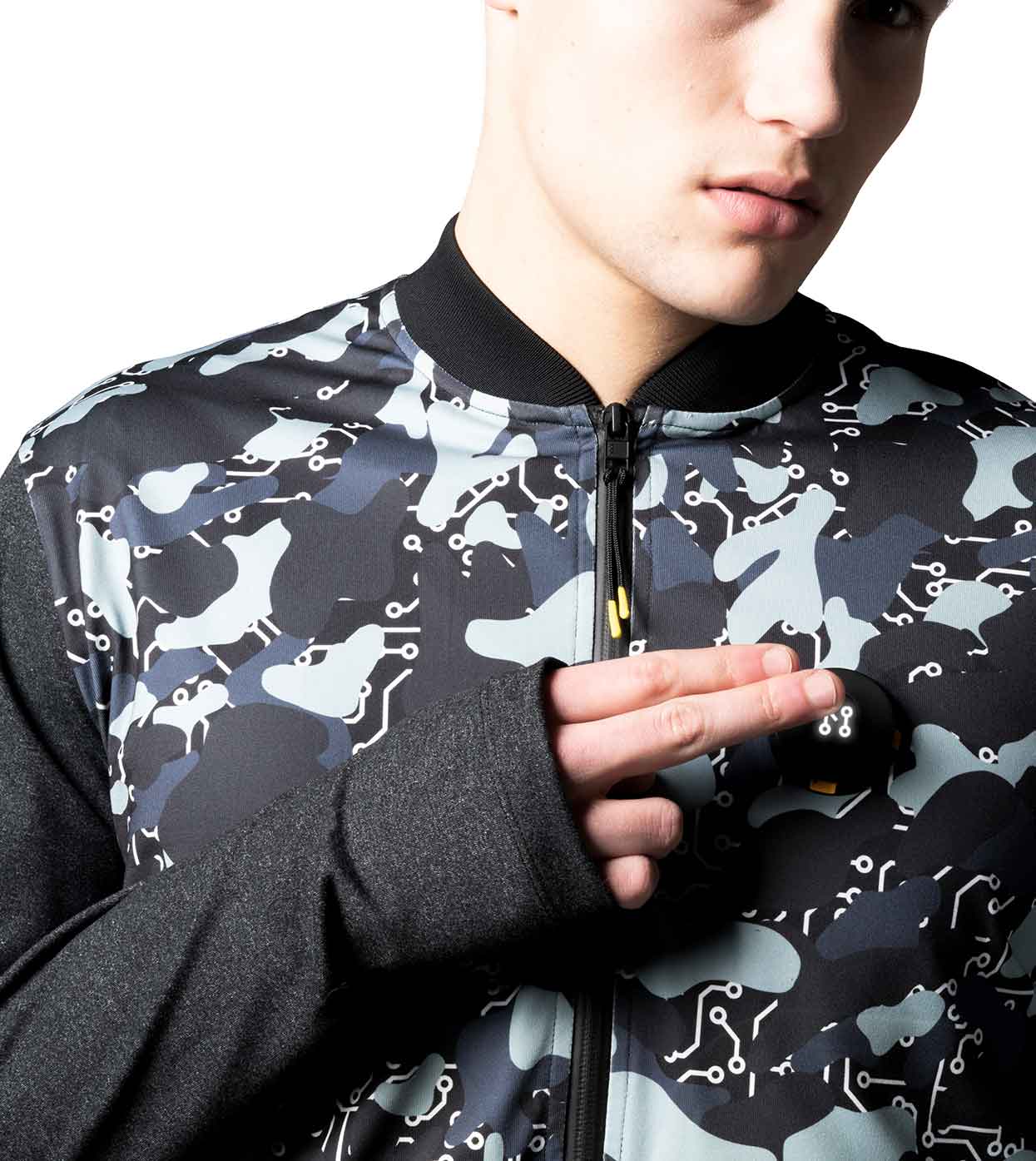 A person is sporting the BLACK-CAMO BOMBER JACKET from Machina - Wearable Technology, featuring an abstract pattern in black, blue, and gray. The jacket has long sleeves and a zipper closure. The individual is holding two small items that look like dice near their chest area.