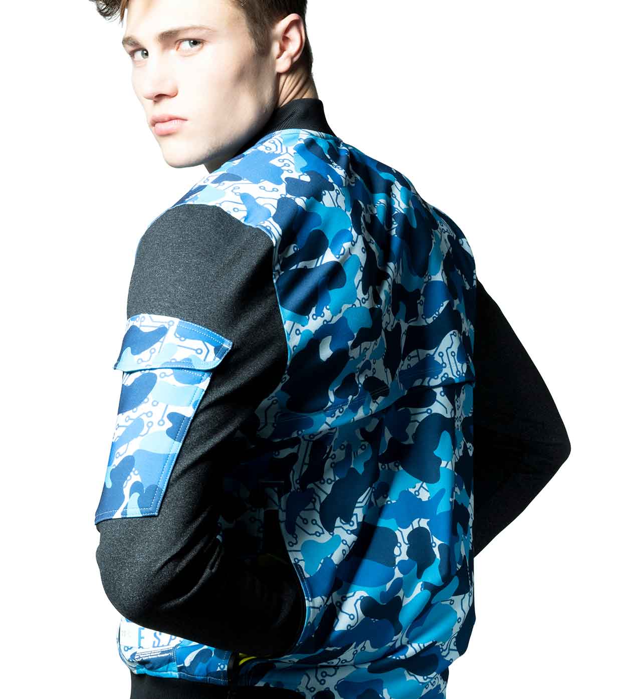 A person dressed in the BLUE-CAMO BOMBER JACKET by Machina - Wearable Technology, featuring black sleeves, stands with their back to the camera, glancing over their shoulder against a white background.