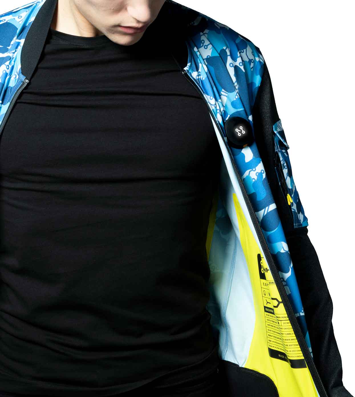 A person dressed in a black shirt is partially hidden by the BLUE-CAMO BOMBER JACKET from Machina - Wearable Technology. They have the jacket open, displaying its vibrant yellow interior. The outer design boasts a camouflage pattern, highlighting cutting-edge fashion tech innovation.