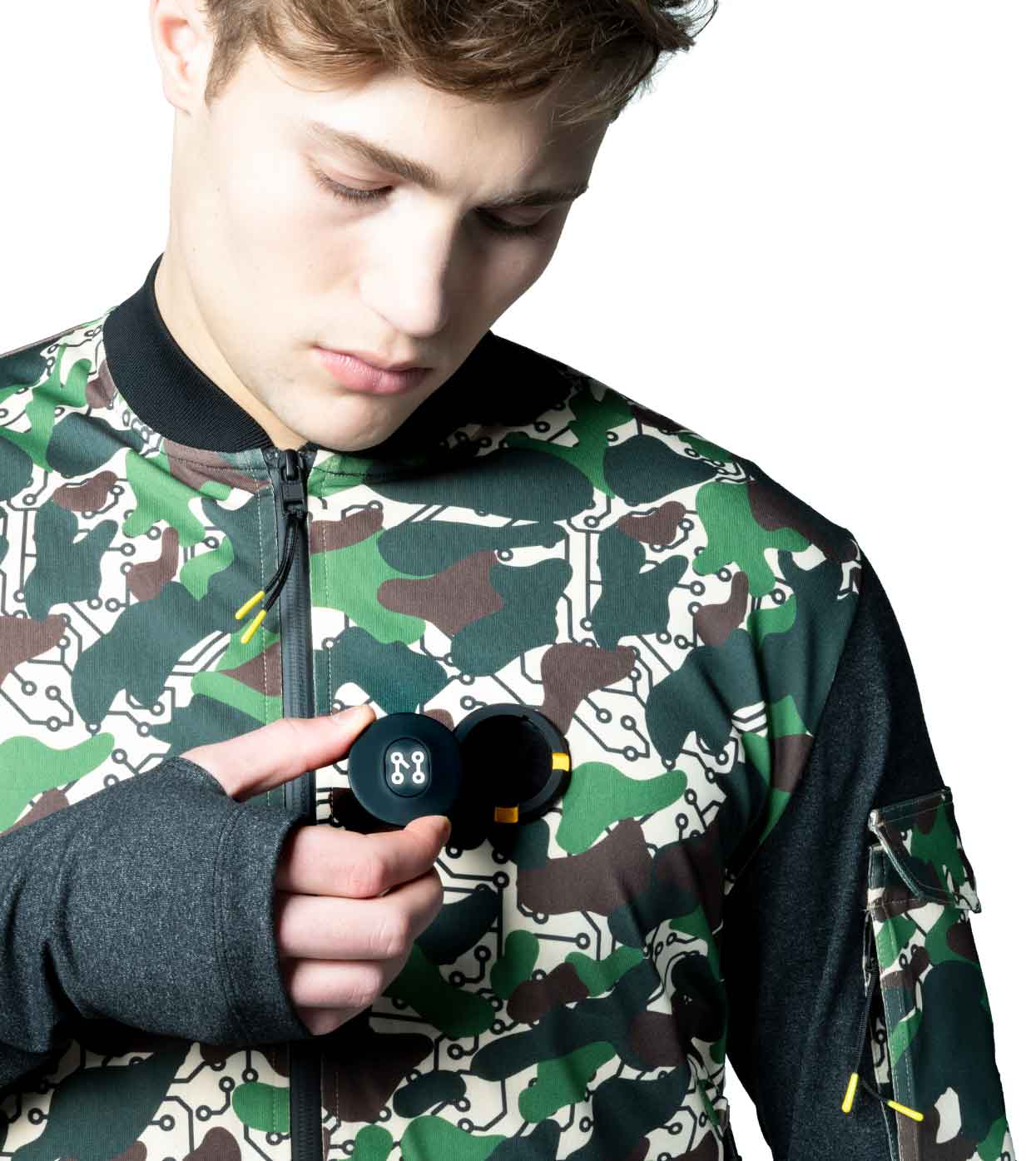 Wearing the DESIERTO CAMO BOMBER JACKET by Machina - Wearable Technology, a person focuses intently on a small black gadget held in their hand. The jacket showcases a camouflage-inspired design with muted greens and browns and is crafted from sustainable fabrics. Their expression is thoughtful against the plain white background.