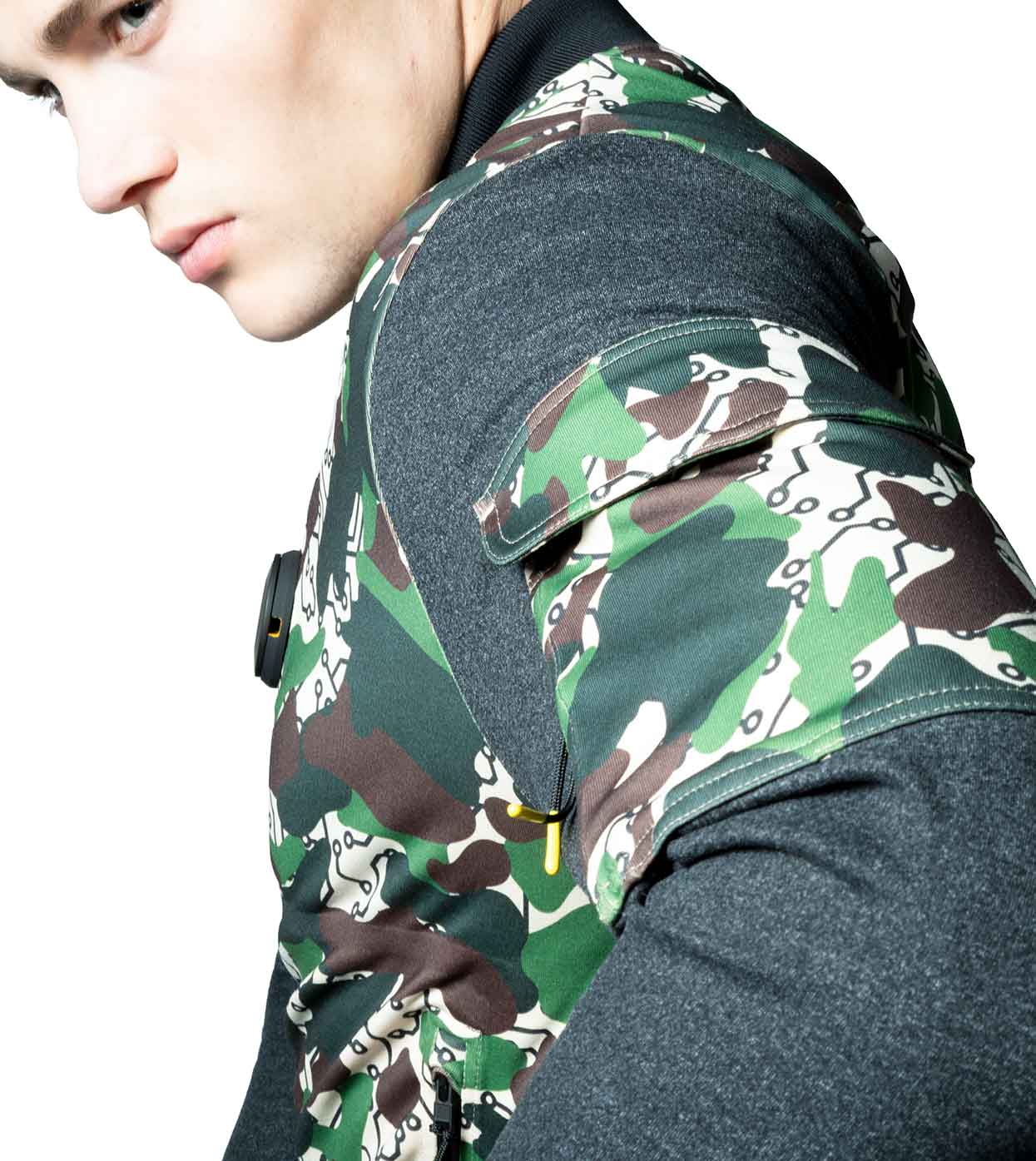 A person is wearing the DESIERTO CAMO BOMBER JACKET by Machina - Wearable Technology over a gray long-sleeve shirt. The image focuses on the upper body, emphasizing the distinctive patchwork design of this fashion-tech piece against a white background.