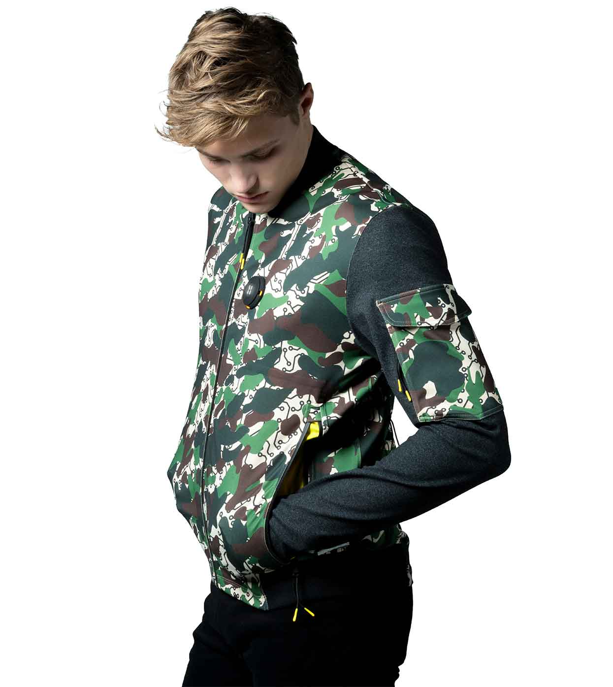 A young man in a sleek DESIERTO CAMO BOMBER JACKET by Machina - Wearable Technology, complete with dark sleeves, looks downward. This eco-friendly jacket is crafted using sustainable fabrics and includes zipper and sleeve pockets. He has short, light brown hair and stands against a plain white background.
