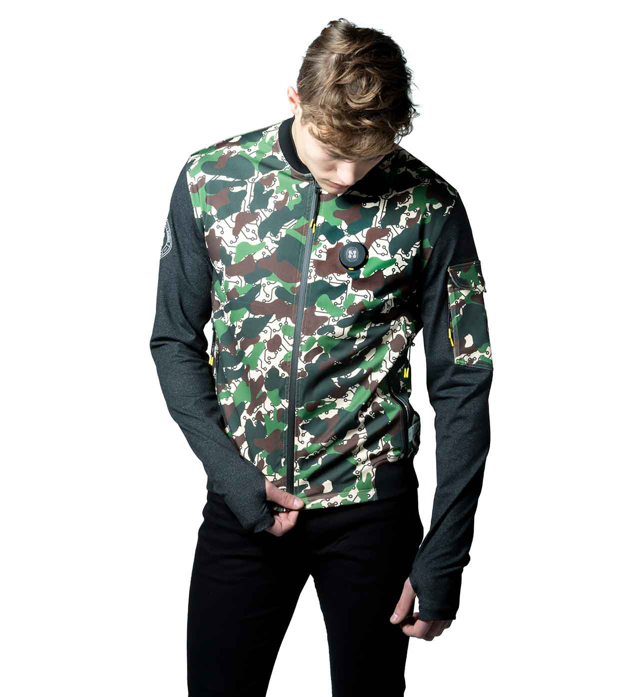 A person wearing the DESIERTO CAMO BOMBER JACKET from Machina - Wearable Technology, which features a camouflage pattern with black sleeves and pants, stands against a white background. The individual's head is slightly bent forward, with one hand partially tucked into the pocket, showcasing modern fashion tech.