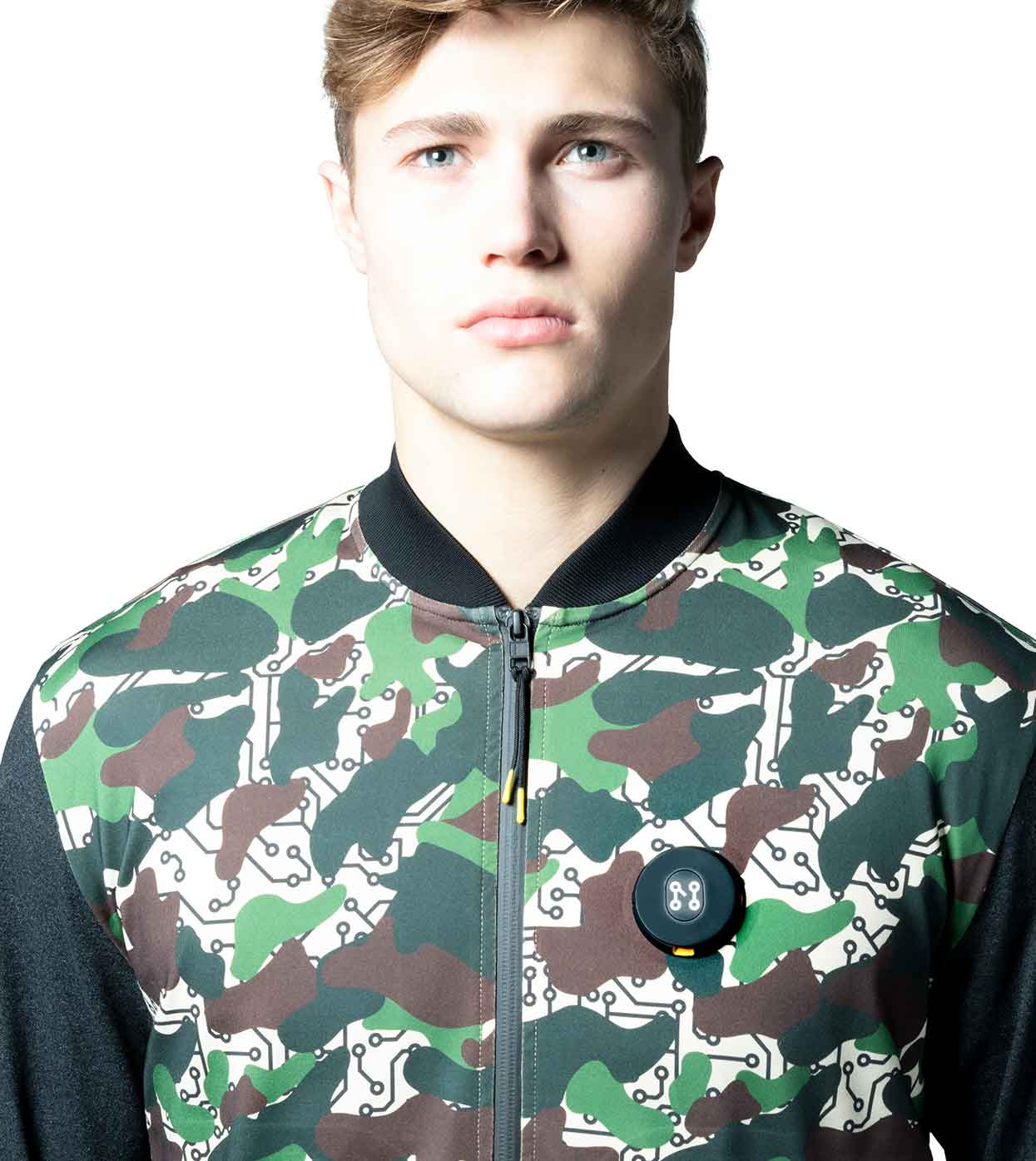 A person with light hair is wearing the Machina - Wearable Technology DESIERTO CAMO BOMBER JACKET, which features a green, brown, and white camouflage pattern and includes a small circular black device. The jacket is crafted from sustainable fabrics, set against a white background.