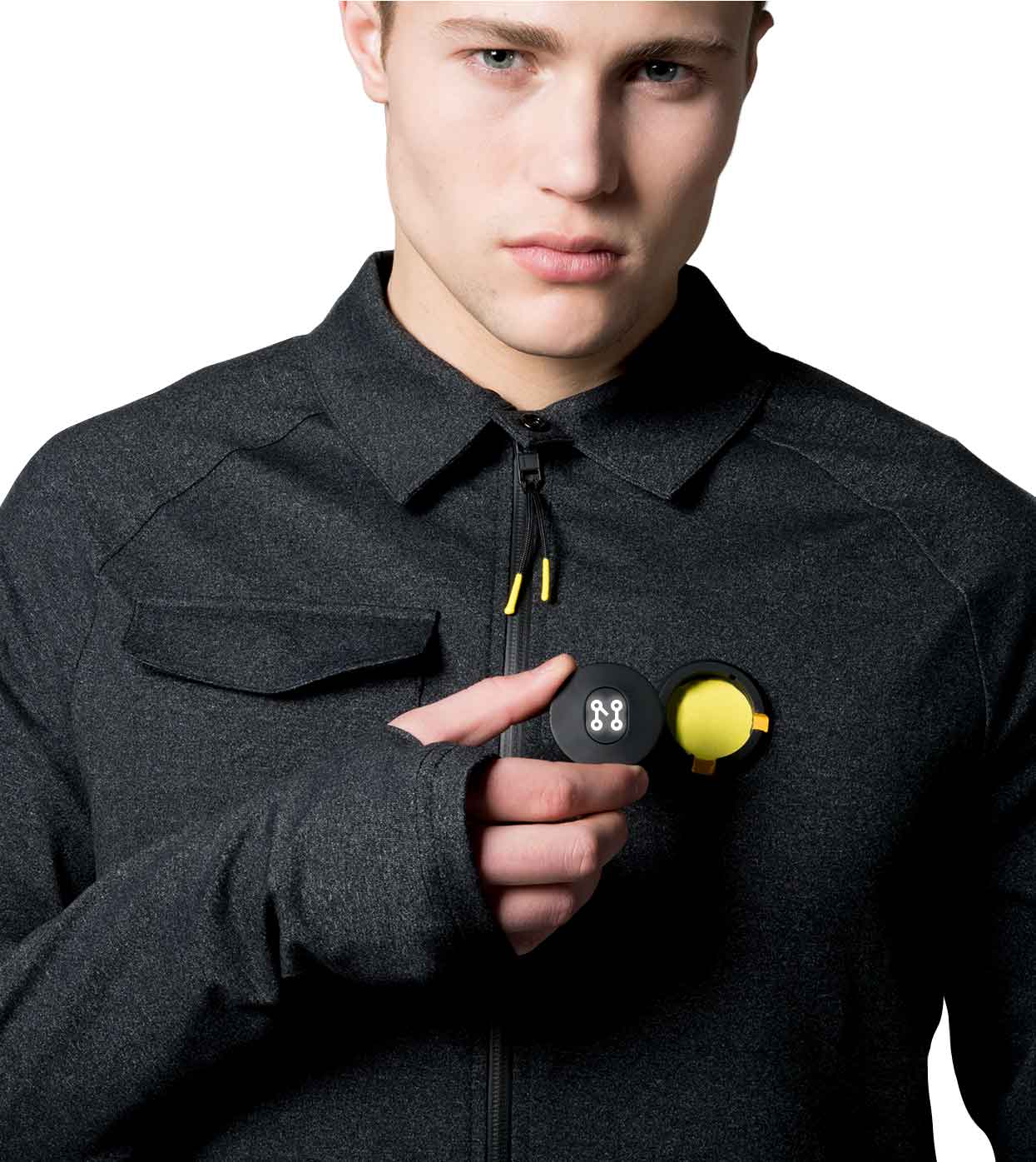 Wearing a BLACK CASUAL JACKET from Machina - Wearable Technology, the person holds a small, round black container with a yellow interior open in one hand. The container is adorned with a white logo on the lid that resembles two connected circles, and the person maintains a neutral expression.