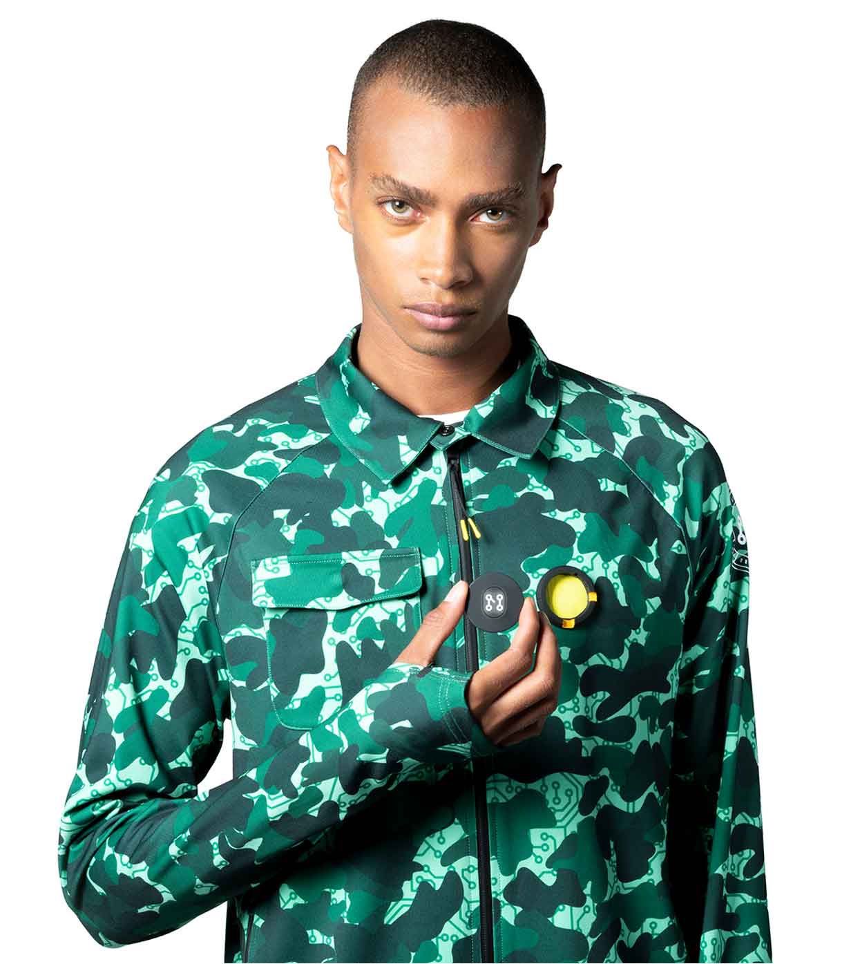 Wearing the AQUA-CAMO CASUAL JACKET by Machina - Wearable Technology, the individual holds a compact, round black gadget with a yellow center against their chest. The plain white backdrop accentuates this subtle yet cutting-edge blend of fashion and technology.