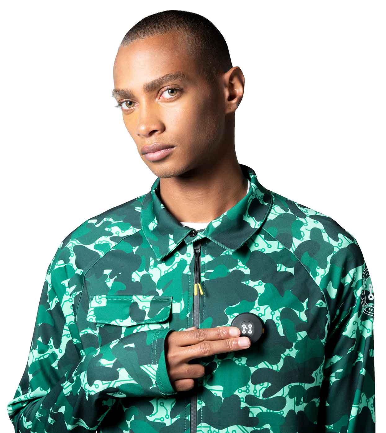 A person with short hair is wearing the AQUA-CAMO CASUAL JACKET from Machina - Wearable Technology, crafted from sustainable fabrics in a striking green and black pattern, while holding a small, round black object featuring a unique design. The plain white background enhances the smart-casual allure of the ensemble.