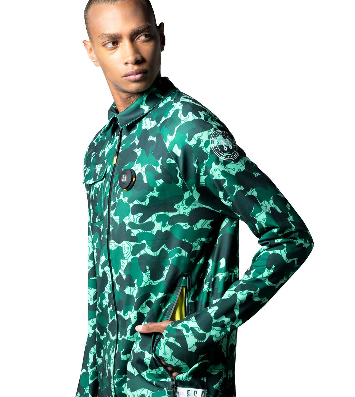 A person poses against a white background wearing the AQUA-CAMO CASUAL JACKET by Machina - Wearable Technology, featuring a smart casual design in green camouflage with large front pockets and a patch on the sleeve, exemplifying fashion technology at its finest.