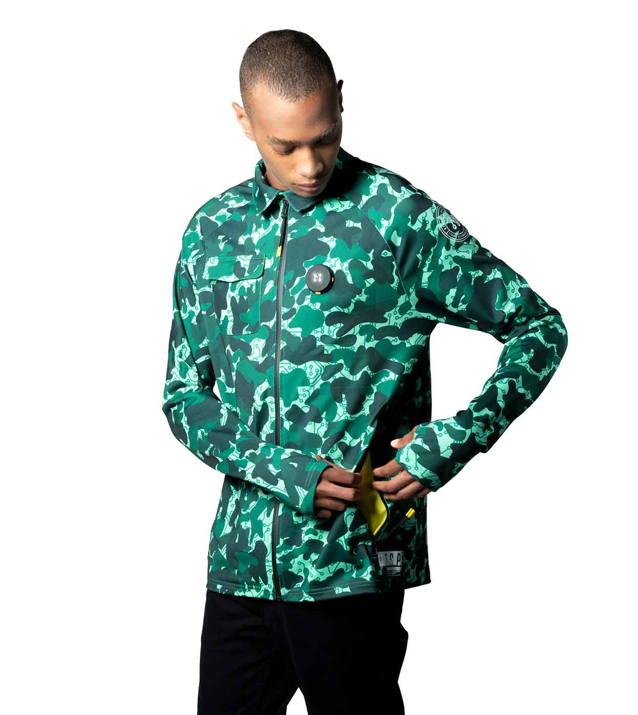 Dressed in the AQUA-CAMO CASUAL JACKET by Machina - Wearable Technology, a person stands against a white background. The jacket, fashioned from sustainable materials and featuring a high collar with zip pockets, is green with a camouflage pattern. They are looking down at the side pocket of their jacket.