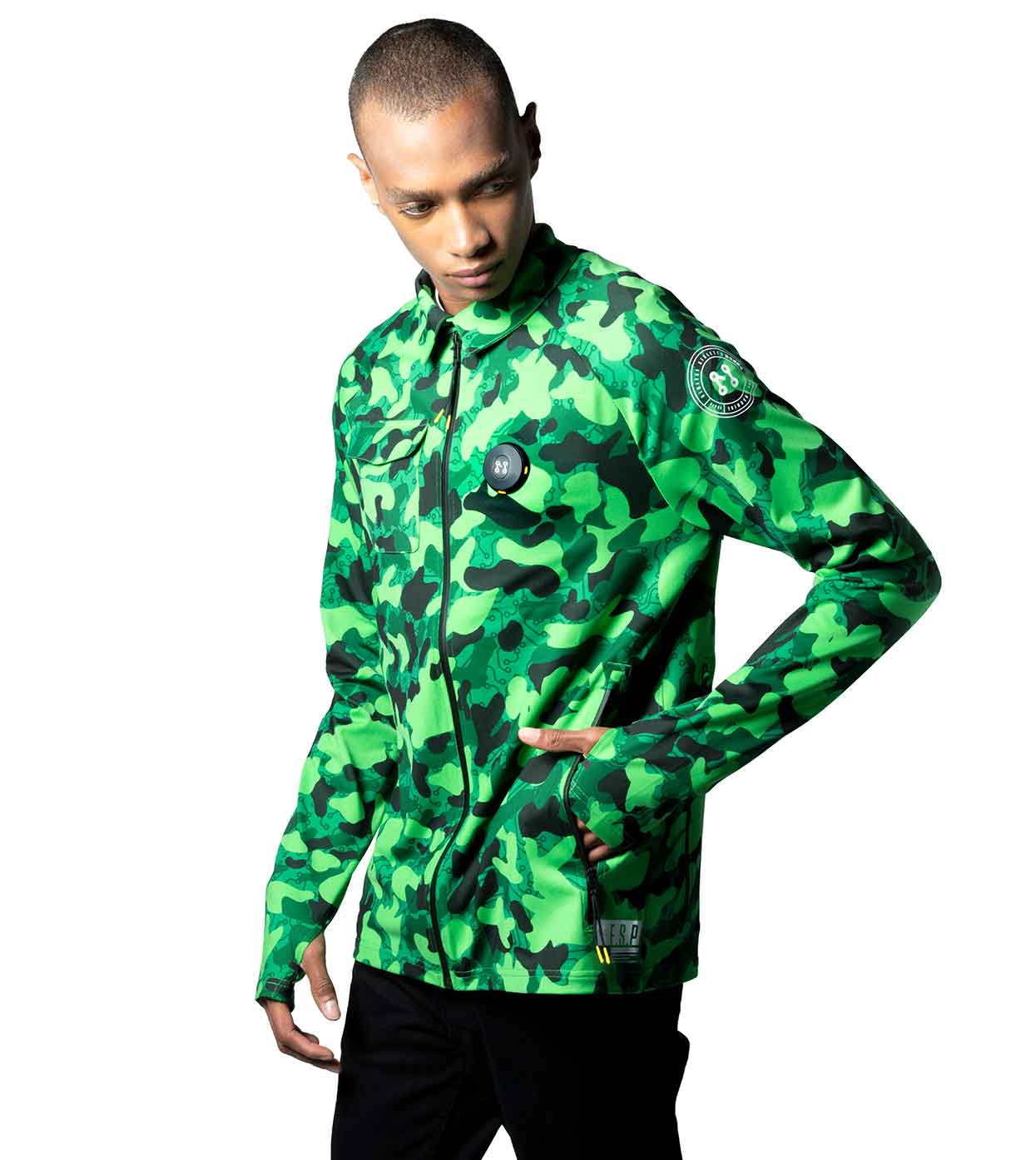 A person dressed in the GREEN-CAMO CASUAL JACKET by Machina - Wearable Technology, crafted from sustainable fabrics and featuring a zipper, stands against a plain white background. This stylish smart casual jacket includes a circular patch and a small emblem on the sleeve. The individual has short hair and pairs the look with black pants.