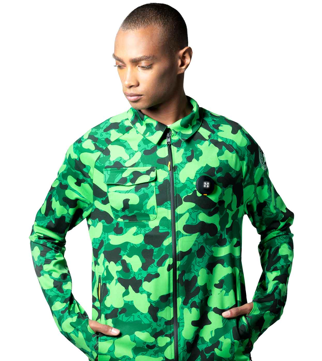 A person wearing the GREEN-CAMO CASUAL JACKET by Machina - Wearable Technology stands with hands comfortably in their pockets, gazing to the side. Crafted from sustainable fabrics, this smart casual jacket includes a distinctive black button or patch near the chest area, set against a plain white background.