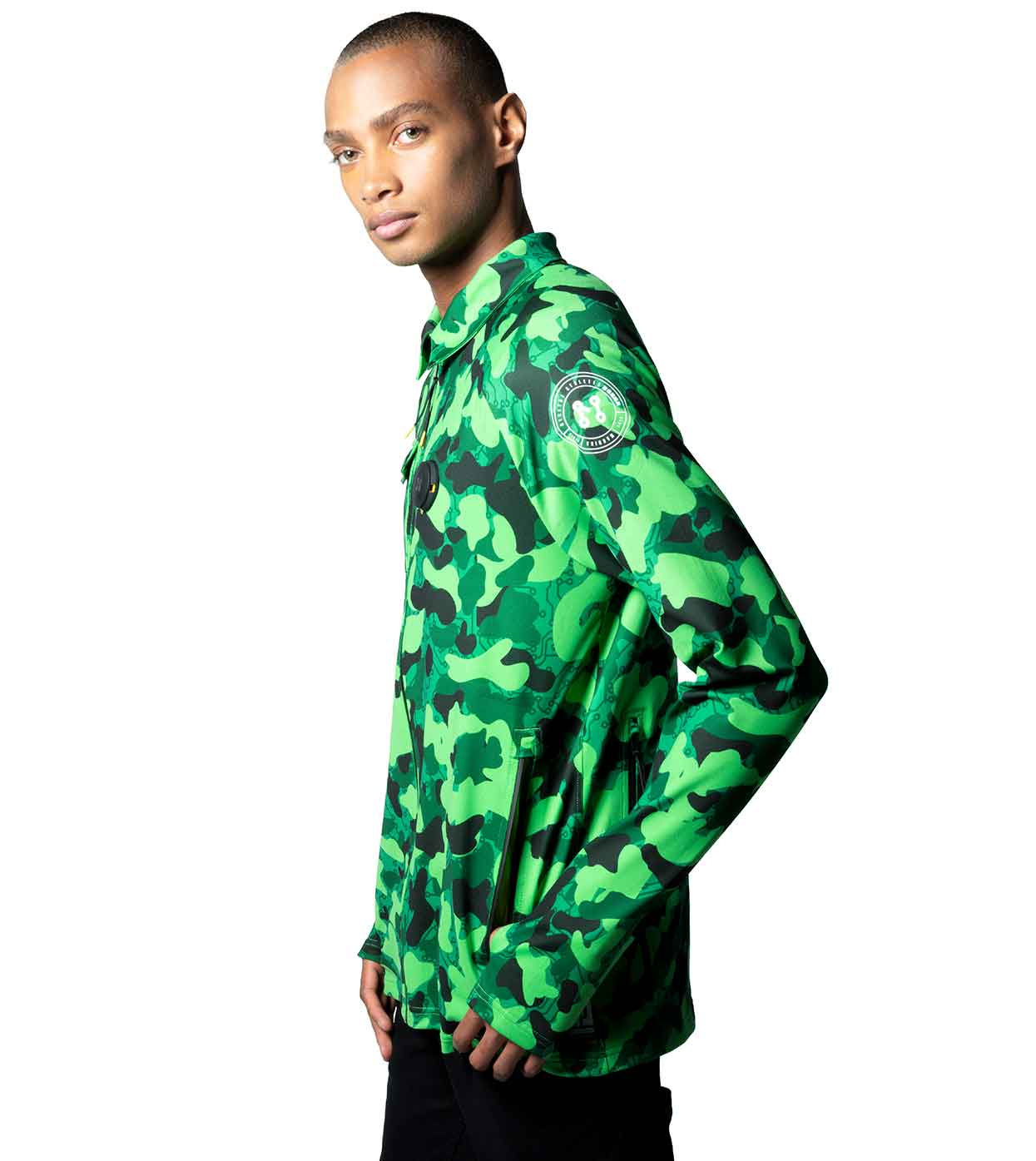 Dressed in the GREEN-CAMO CASUAL JACKET by Machina - Wearable Technology, an individual poses against a white background, glancing over their shoulder. The jacket is crafted from sustainable fabrics and showcases a circular emblem on the sleeve, combining stylish design with eco-friendly principles.