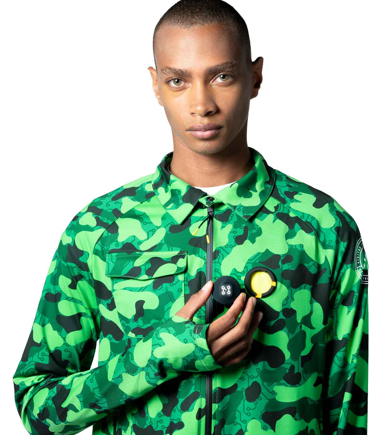A person wearing the GREEN-CAMO CASUAL JACKET from Machina - Wearable Technology holds two small items, one black and one yellow, in their hand against a white background.