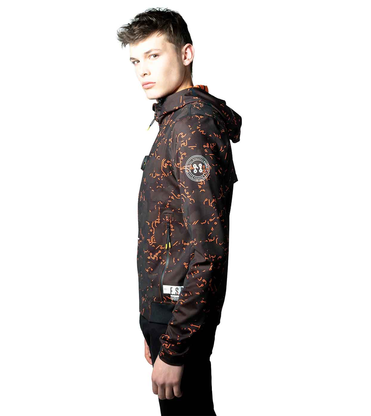 An individual wearing the ELECTRIC ORANGE HOODIE by Machina - Wearable Technology, designed with black fabric, vibrant orange patterns, and waterproof zippers, poses against a white background while gazing sideways. The innovative fashion tech hoodie also showcases a circular patch on the shoulder.