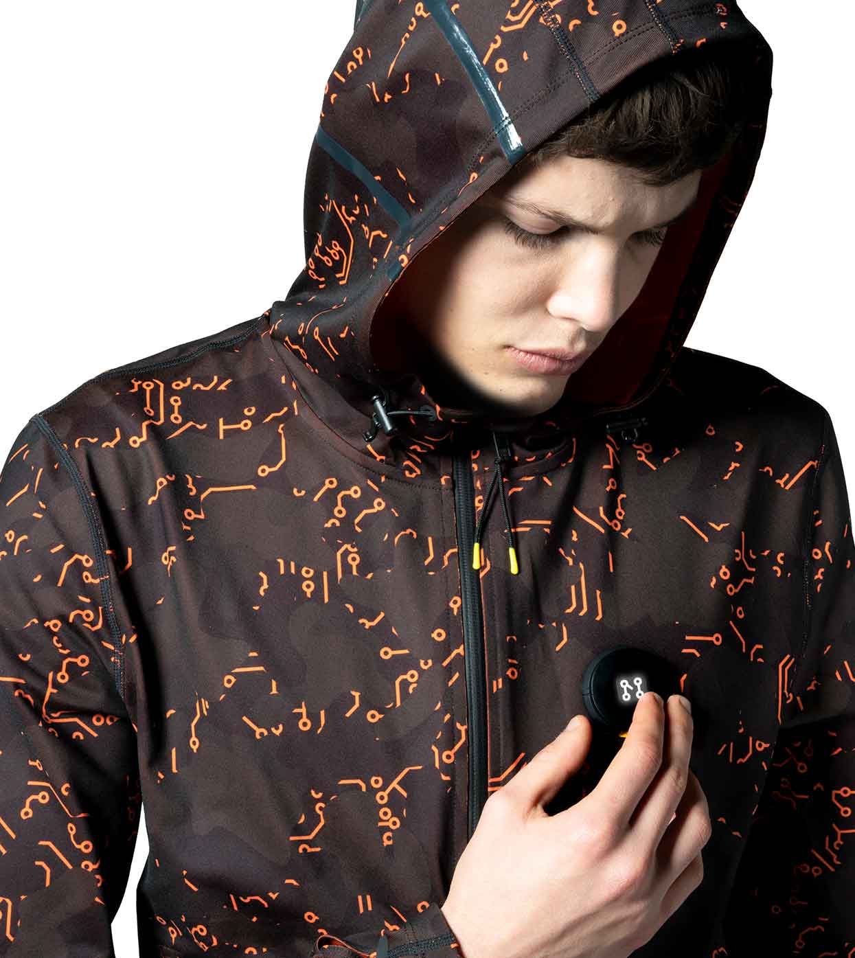 A person dressed in the ELECTRIC ORANGE HOODIE by Machina - Wearable Technology, featuring an orange circuit pattern, is observing a small black device they are holding. The hood is up and includes waterproof zippers, set against a plain white background.
