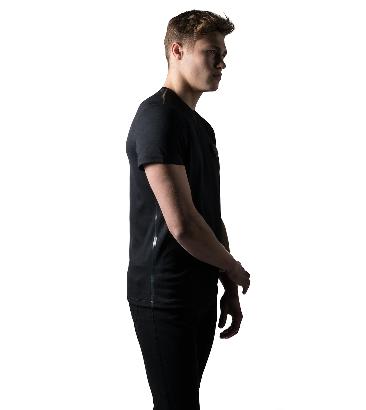A person in Machina - Wearable Technology's Smart Black T-shirt and matching pants, made from sustainable fabrics, stands against a white backdrop with a slight side-facing pose. The lighting casts a dramatic shadow effect.