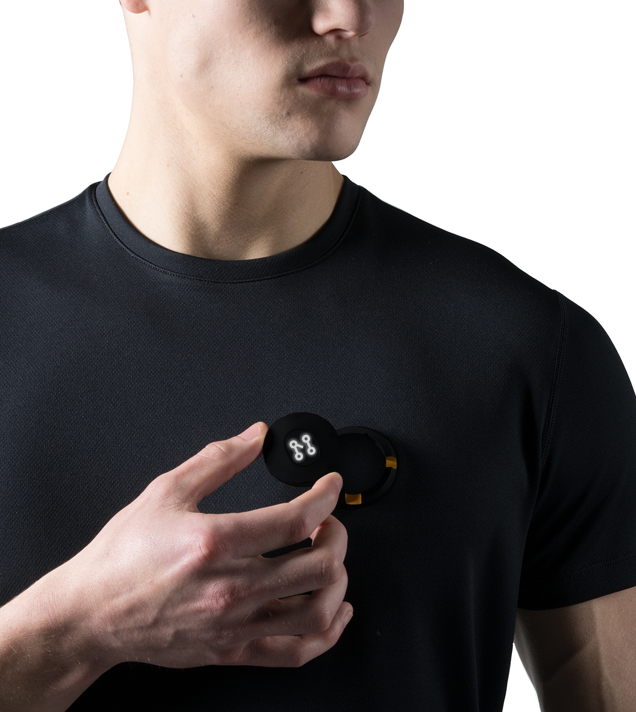 A wearer of the Machina - Wearable Technology's Smart Black T-shirt places a small, round device adorned with a white emblem on their chest. In their hand is the open case, displaying the contents. The scene unfolds against a white backdrop, partially revealing the face—a subtle nod to cutting-edge smart T-shirt integration.