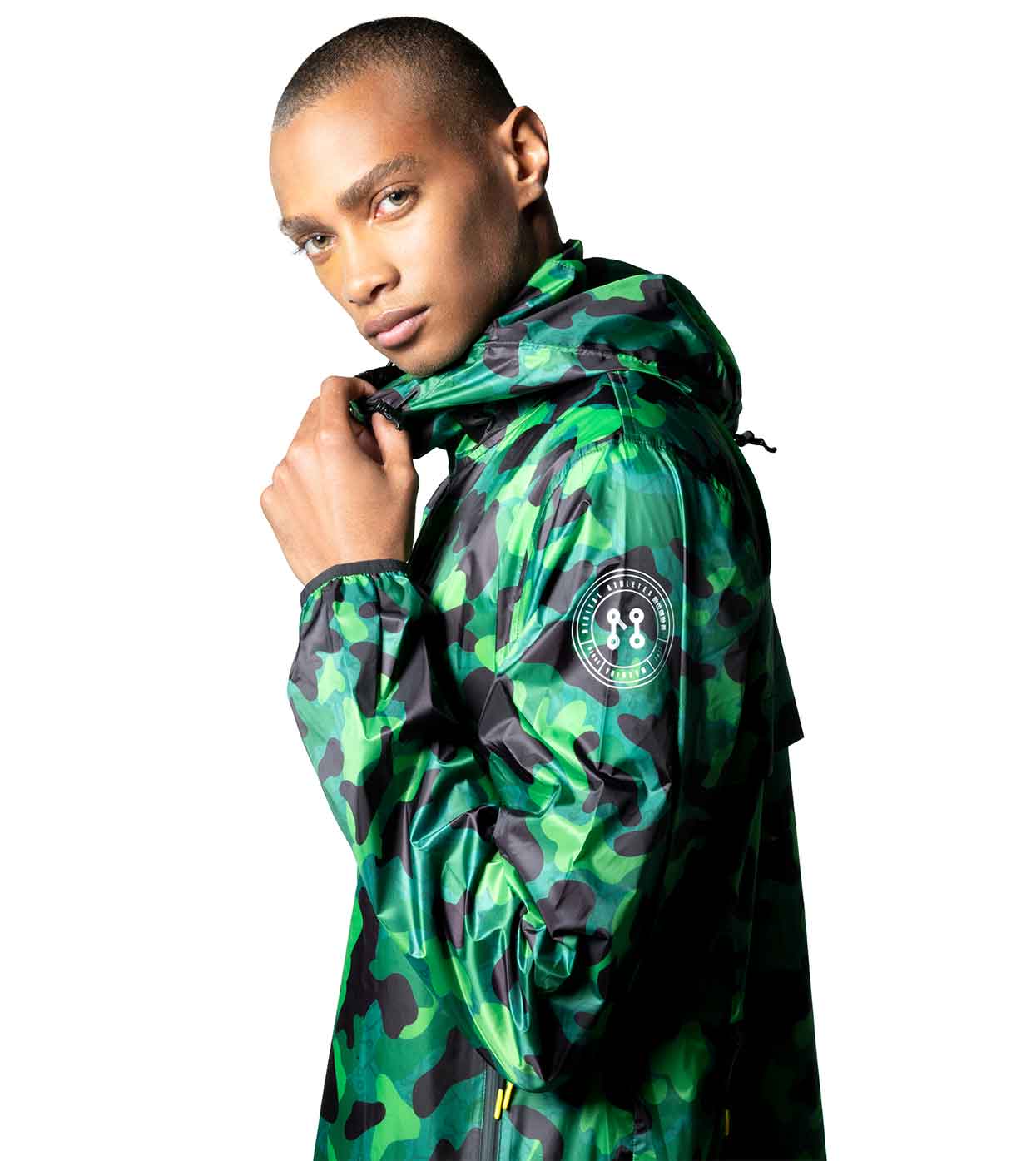 A person is wearing the AQUA-CAMO WINDBREAKER by Machina - Wearable Technology. This green aqua camouflage jacket with a hood features a circular patch design on the sleeve and showcases advanced fashion technology. The person is looking towards the camera, with one hand touching the jacket's collar, set against a plain white background.
