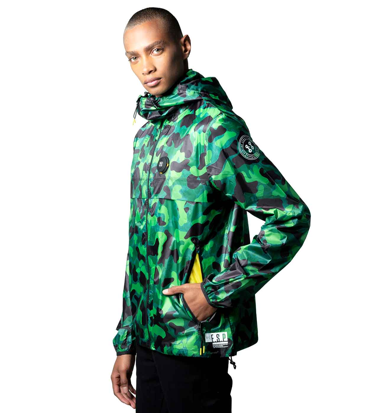 An individual dressed in Machina - Wearable Technology's AQUA-CAMO WINDBREAKER, featuring tech-enhanced patch details on the sleeves and chest, along with black pants, is seen glancing back over their shoulder against a plain white background. The jacket includes a hood and showcases an aqua camouflage design.