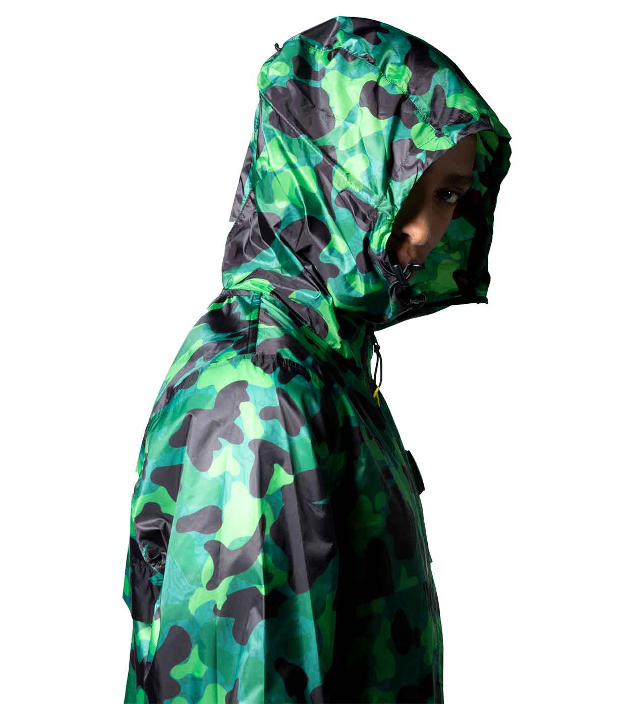 The image features an individual in profile wearing the AQUA-CAMO WINDBREAKER by Machina - Wearable Technology. The hood conceals most of their head, and their face is partially visible in shadow against a white background, highlighting the blend of fashion and technology in this cutting-edge design.