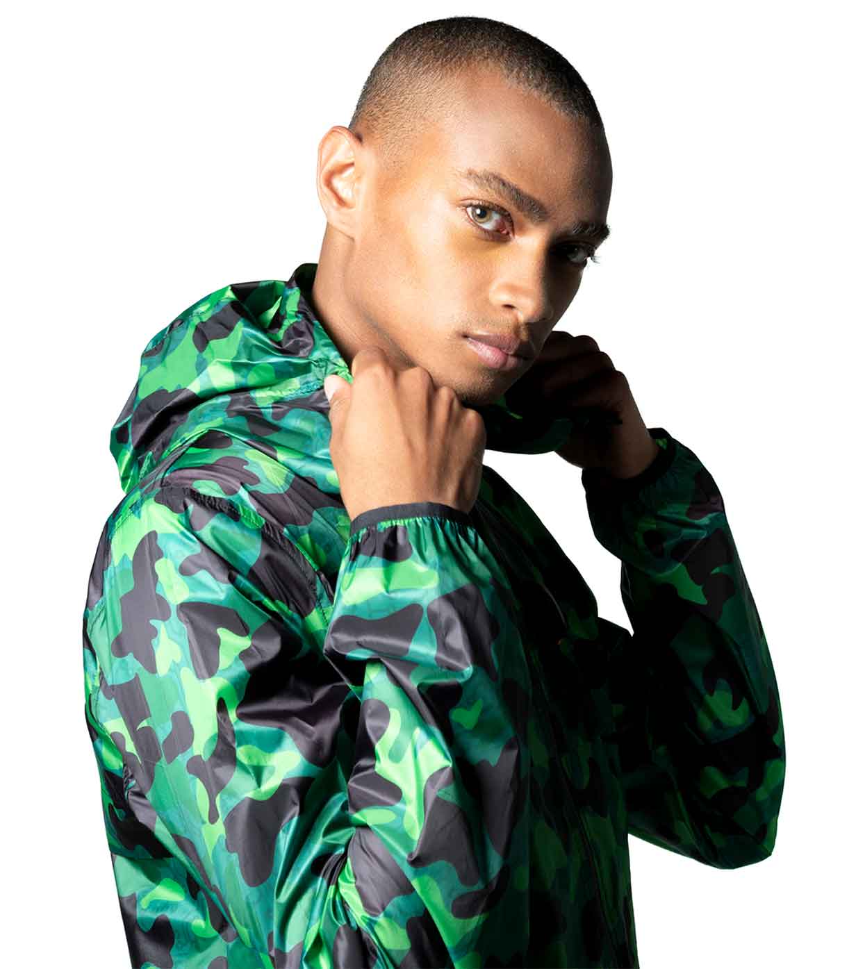 Wearing the AQUA-CAMO WINDBREAKER by Machina - Wearable Technology, a person dressed in a shiny green and black camouflage jacket holds the collar while looking directly at the camera. The hood of the smart windbreaker is up against a plain white background.