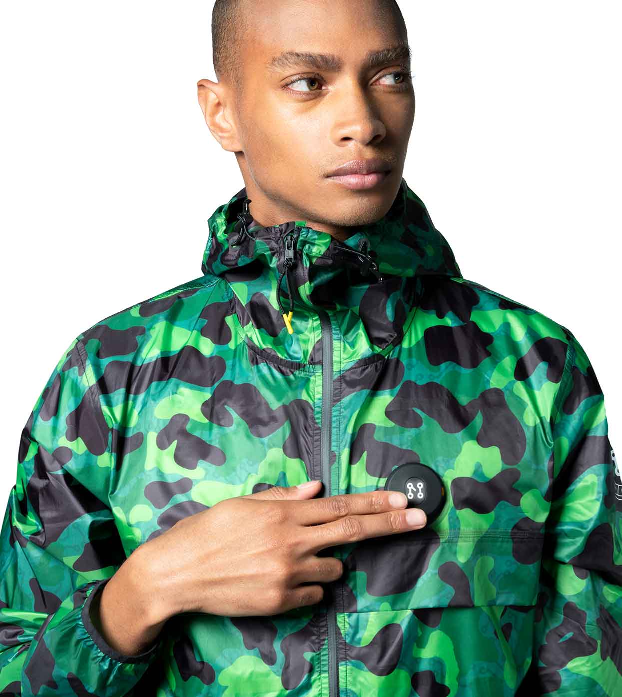 Dressed in the AQUA-CAMO WINDBREAKER by Machina - Wearable Technology, a person embodies modern style and innovation. They wear the green camouflage jacket with a hood and highlight its advanced fashion technology by pointing two fingers towards the circular chest patch against a plain white background.
